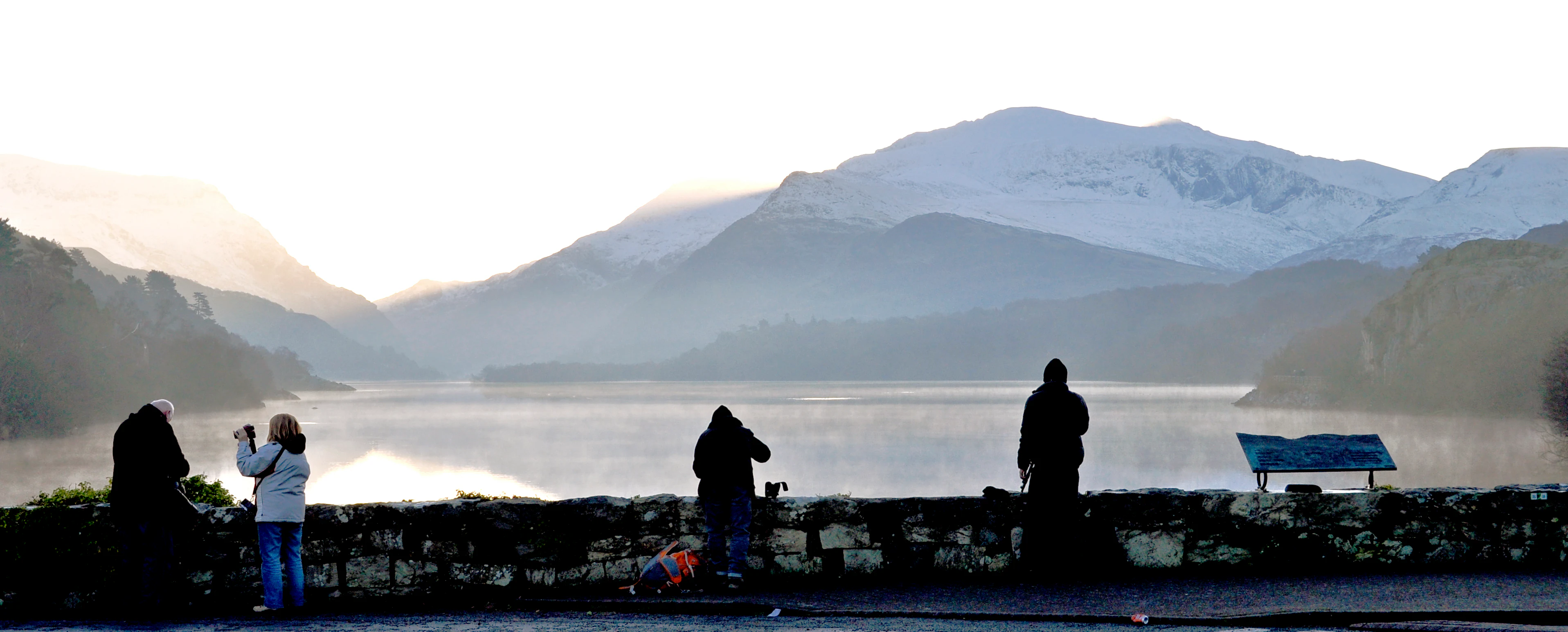 Silhouettes of four people standing by a stone wall overlooking a lake with snow-capped mountains in the background during sunrise