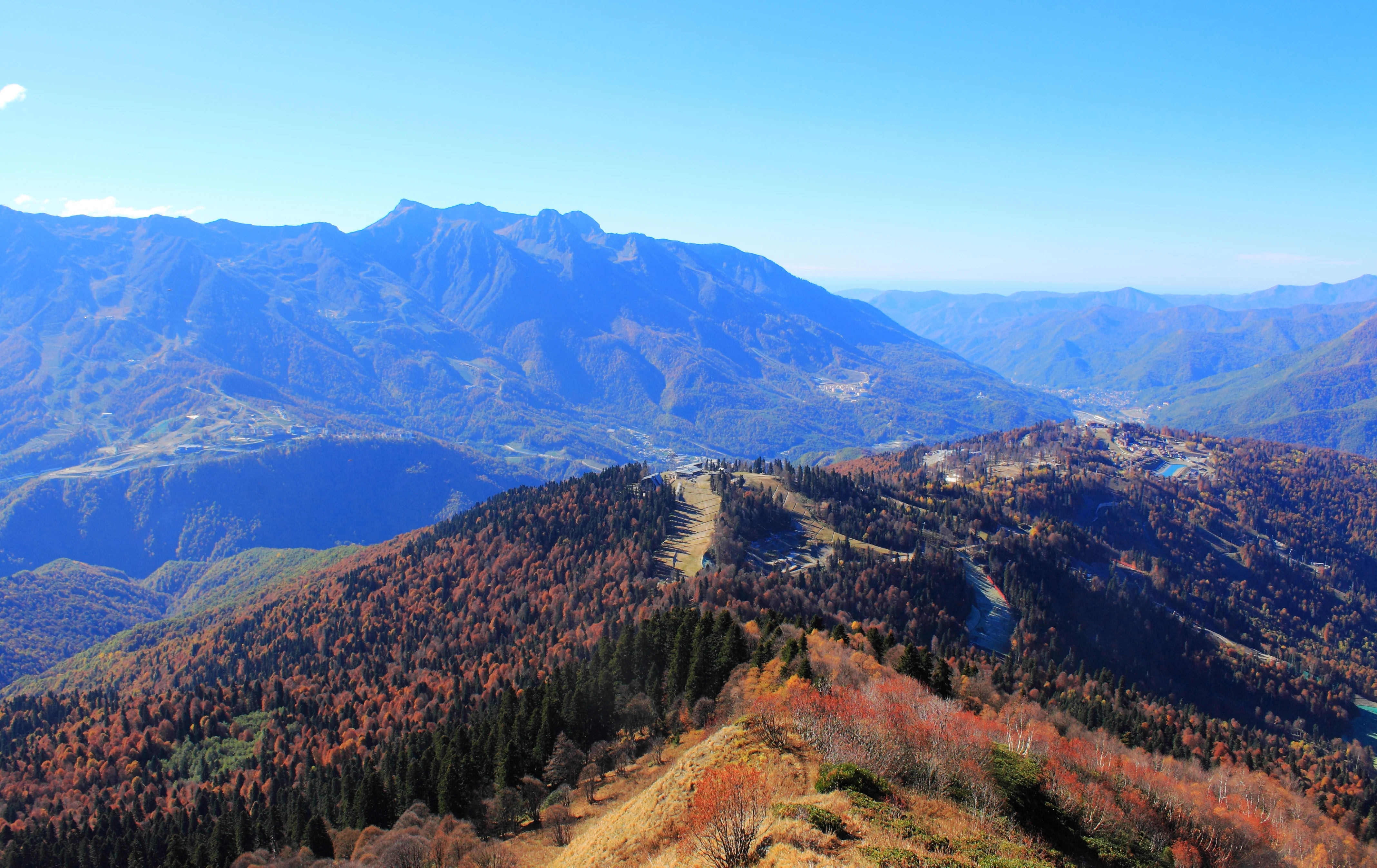 Una vista panorámica en ángulo alto de laderas montañosas onduladas cubiertas de bosque otoñal bajo un cielo azul despejado, con líneas de crestas visibles y terreno de valles.