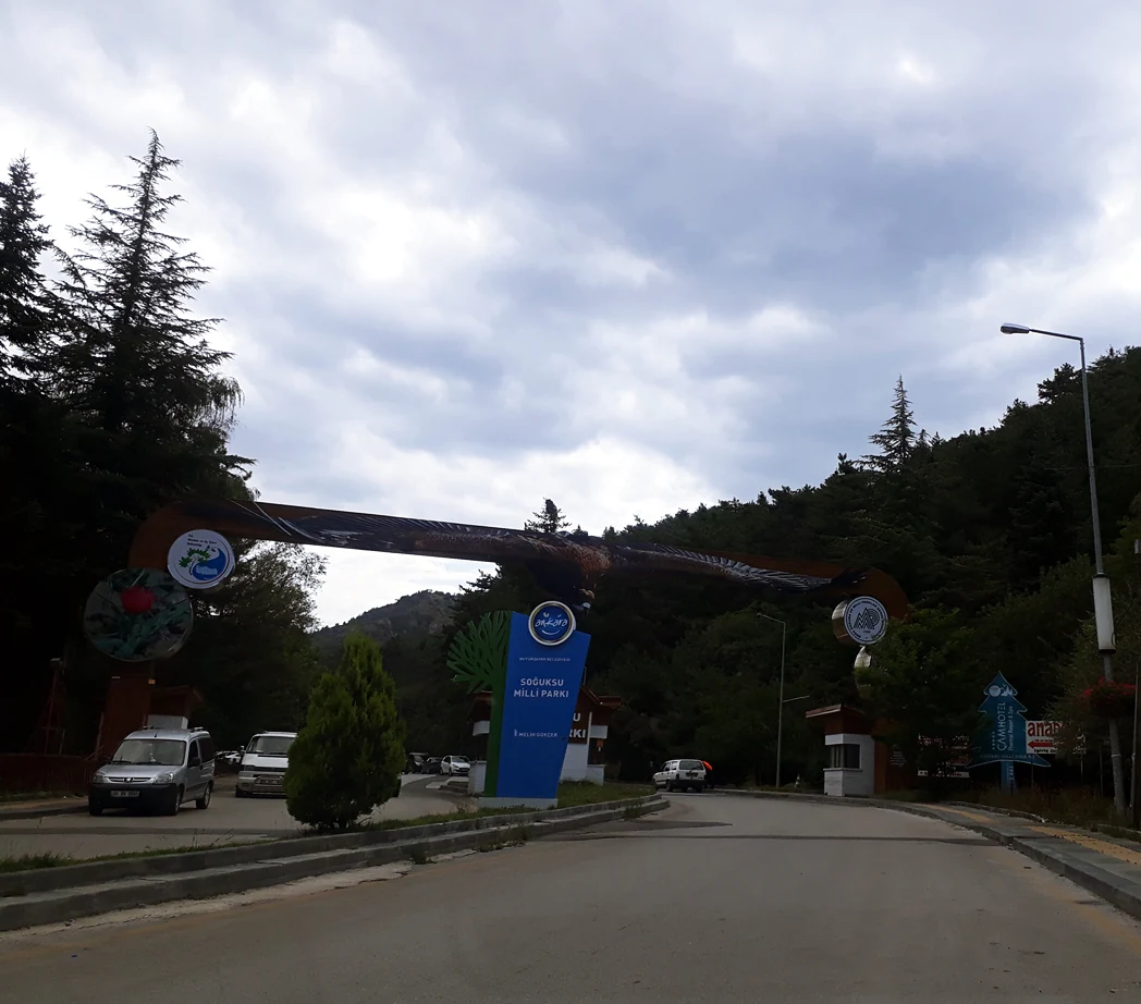 Large archway entrance sign for Soğuksu National Park, blue informational sign, vehicles on paved road, forested hills and trees in background