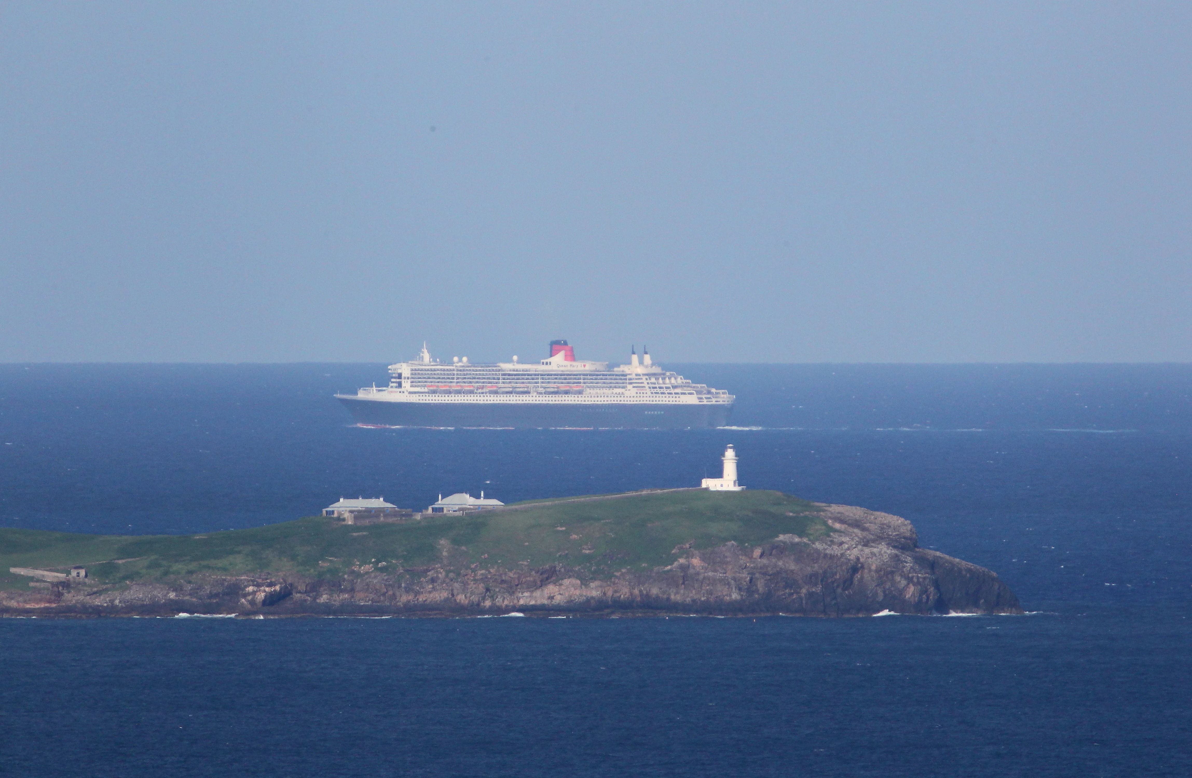 Un gran crucero navegando en mar abierto detrás de una isla verde coronada por un faro blanco.