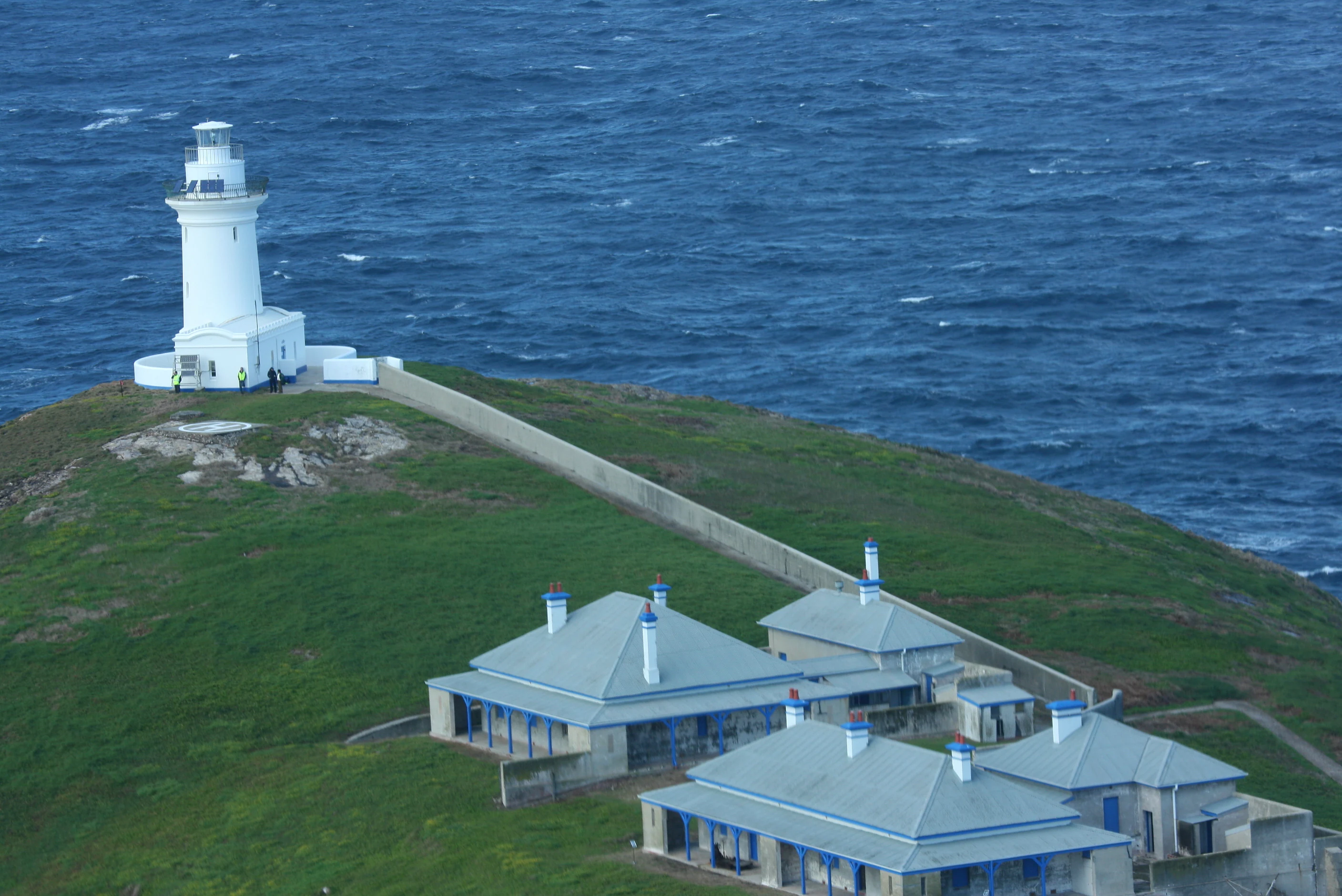 Una vista aérea del faro blanco de South Solitary Island y varias cabañas adyacentes de los cuidadores con techos grises en una ladera cubierta de hierba de la isla, con el profundo océano azul al fondo.