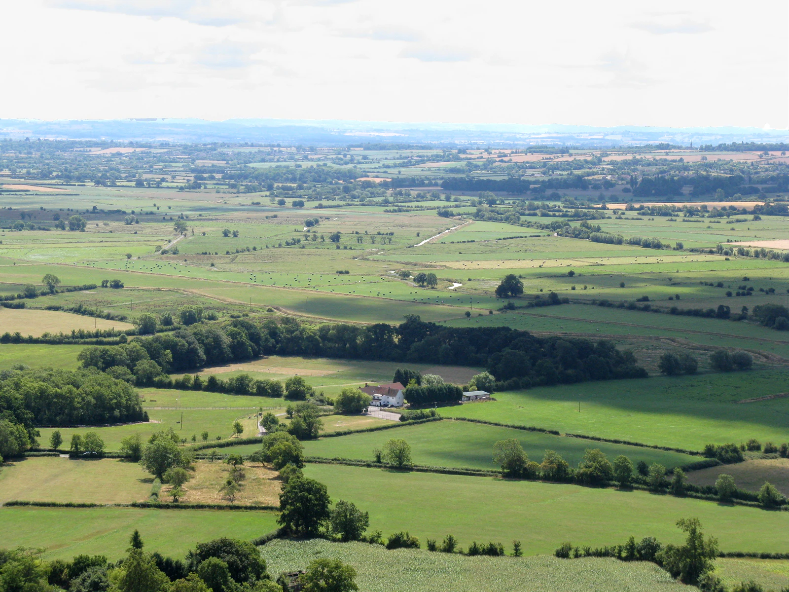 Una vista elevada y de gran angular de los Somerset Levels, que muestra un mosaico rural plano y verde de pastos, campos y setos que se extienden hasta un horizonte lejano bajo un cielo nublado.