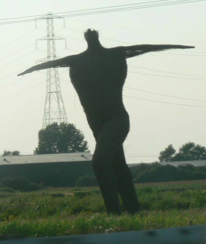 Una escultura grande y oscura con forma humana, hecha de ramas de sauce tejidas, de pie en un campo con los brazos extendidos contra un cielo pálido y una torre eléctrica al fondo.