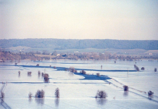 Una vista amplia, brumosa y en ángulo alto de marismas y campos inundados en Southlake Moor en invierno, con árboles desnudos dispersos y una cresta baja distante.