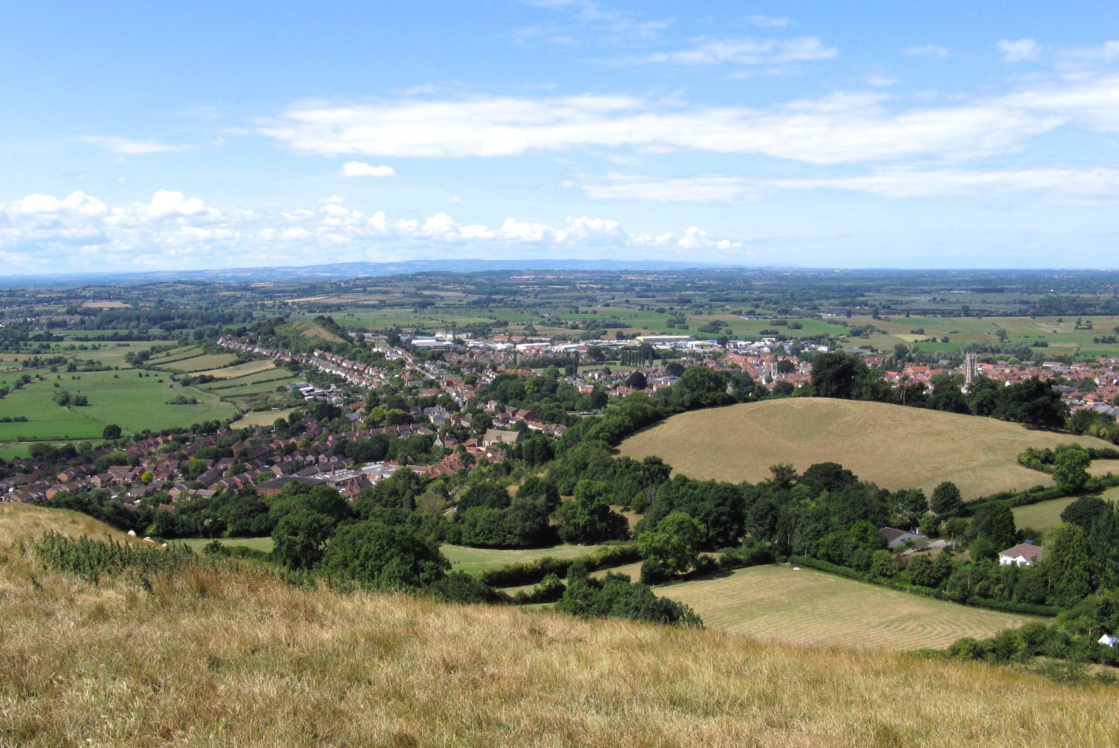 Una vista elevada y de gran angular desde una colina cubierta de hierba que domina la localidad de Glastonbury, rodeada de campos verdes y las extensas y llanas Somerset Levels bajo un cielo azul.