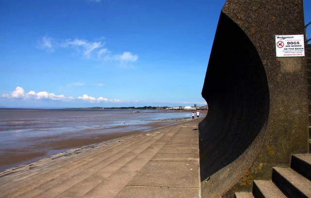 Una vista amplia de un muro de hormigón y escalones que dan a una vasta playa de fango intermareal bajo un cielo azul despejado, con una oscura característica arquitectónica curva en primer plano.