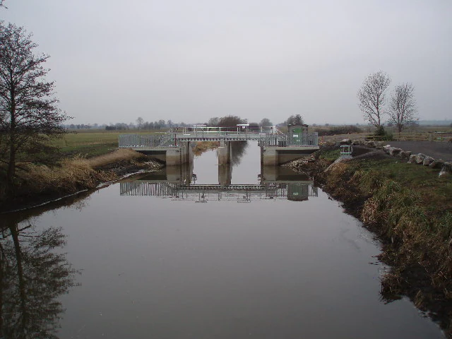 Un canal tranquilo y estrecho fluye hacia una estructura de compuerta de hormigón en un paisaje de humedal plano bajo un cielo nublado.