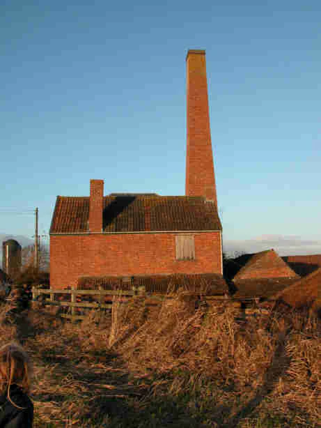Un edificio histórico de ladrillo rojo con una chimenea alta se alza en un campo al atardecer.