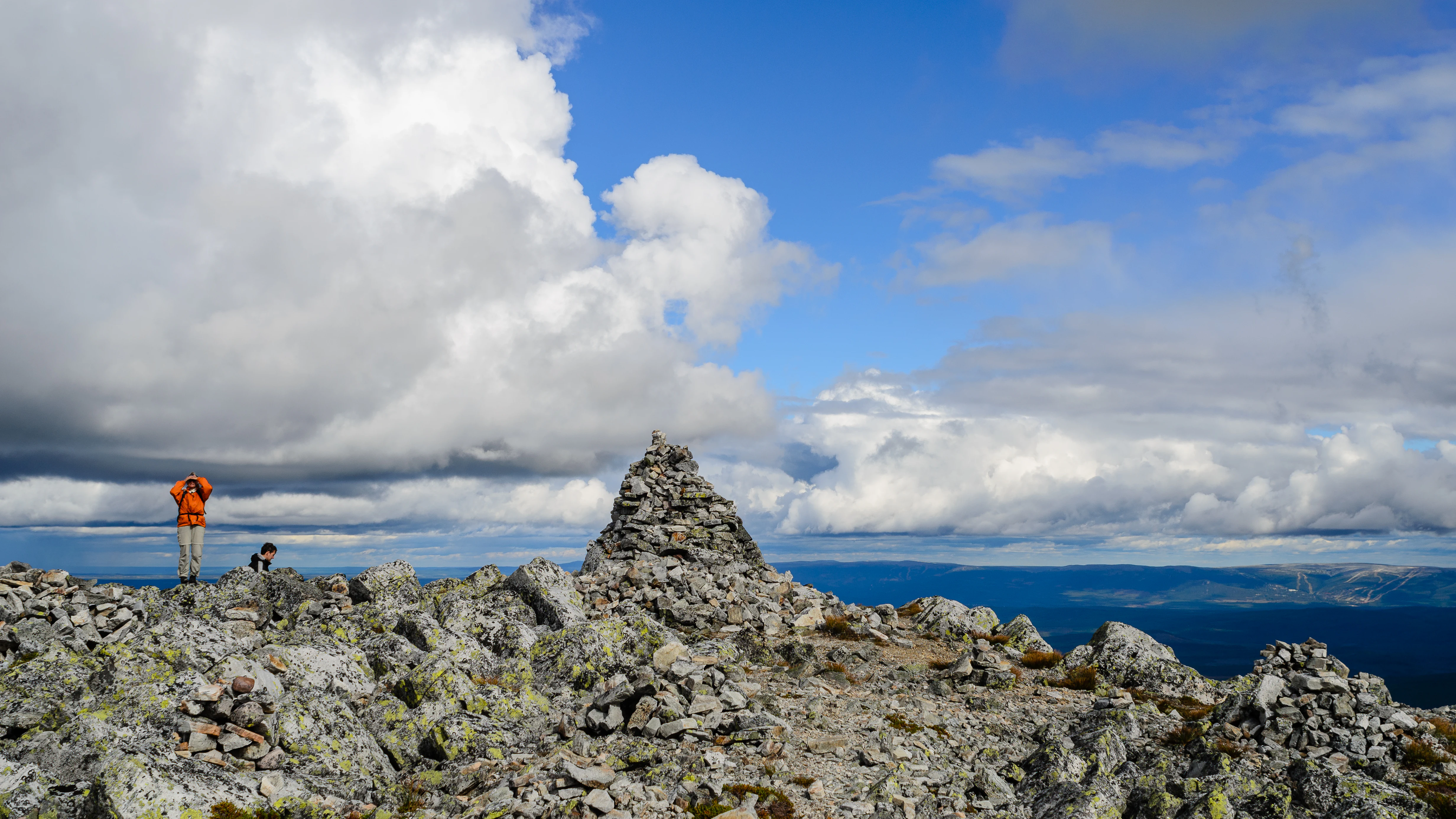 Rocky mountain summit featuring a stone cairn, two small figures of people, and a cloudy sky