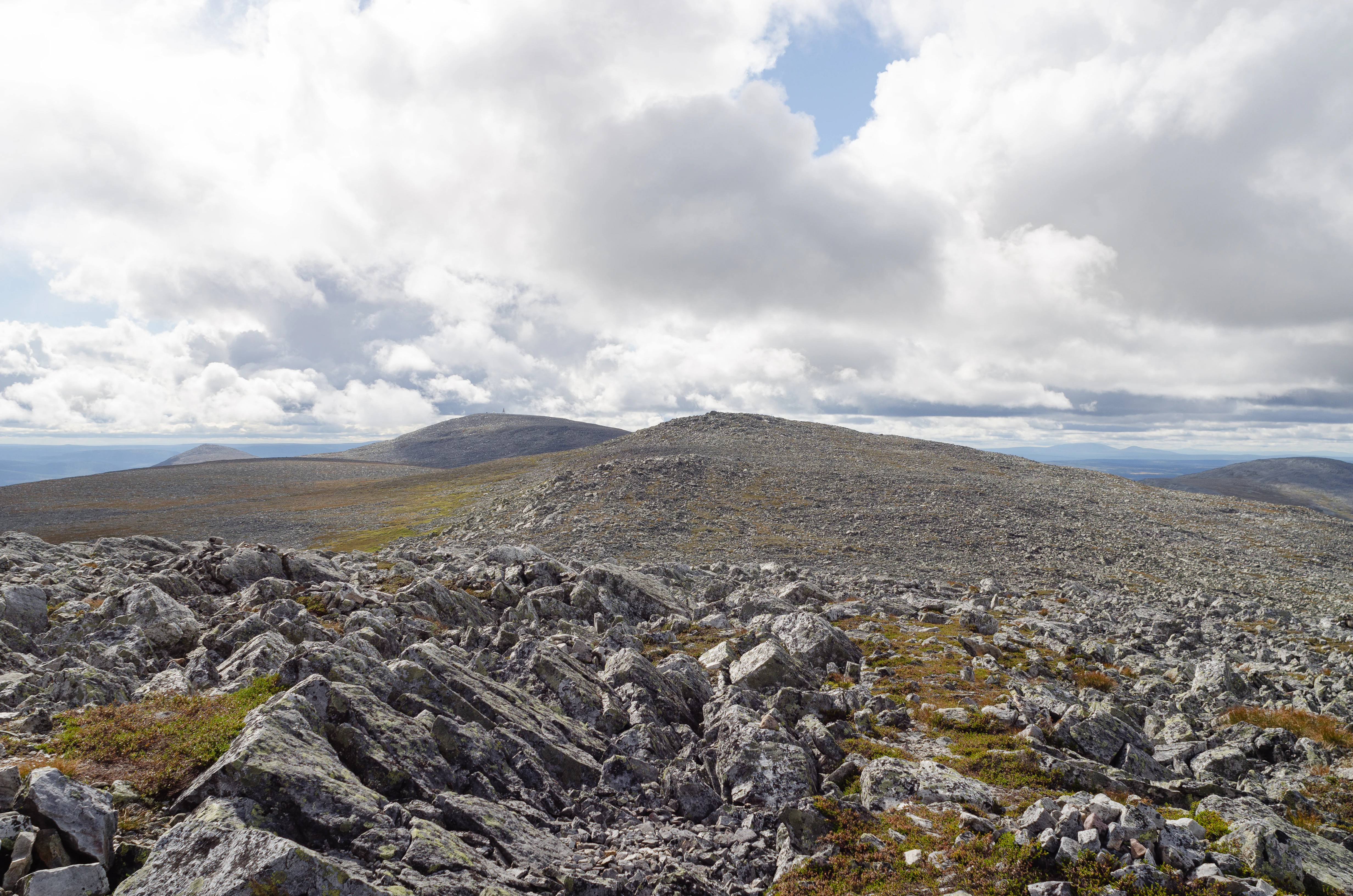 Rocky terrain with patches of moss and lichen in the foreground, extending to a flat-topped mountain summit under a partly cloudy sky with distant mountains visible on the horizon