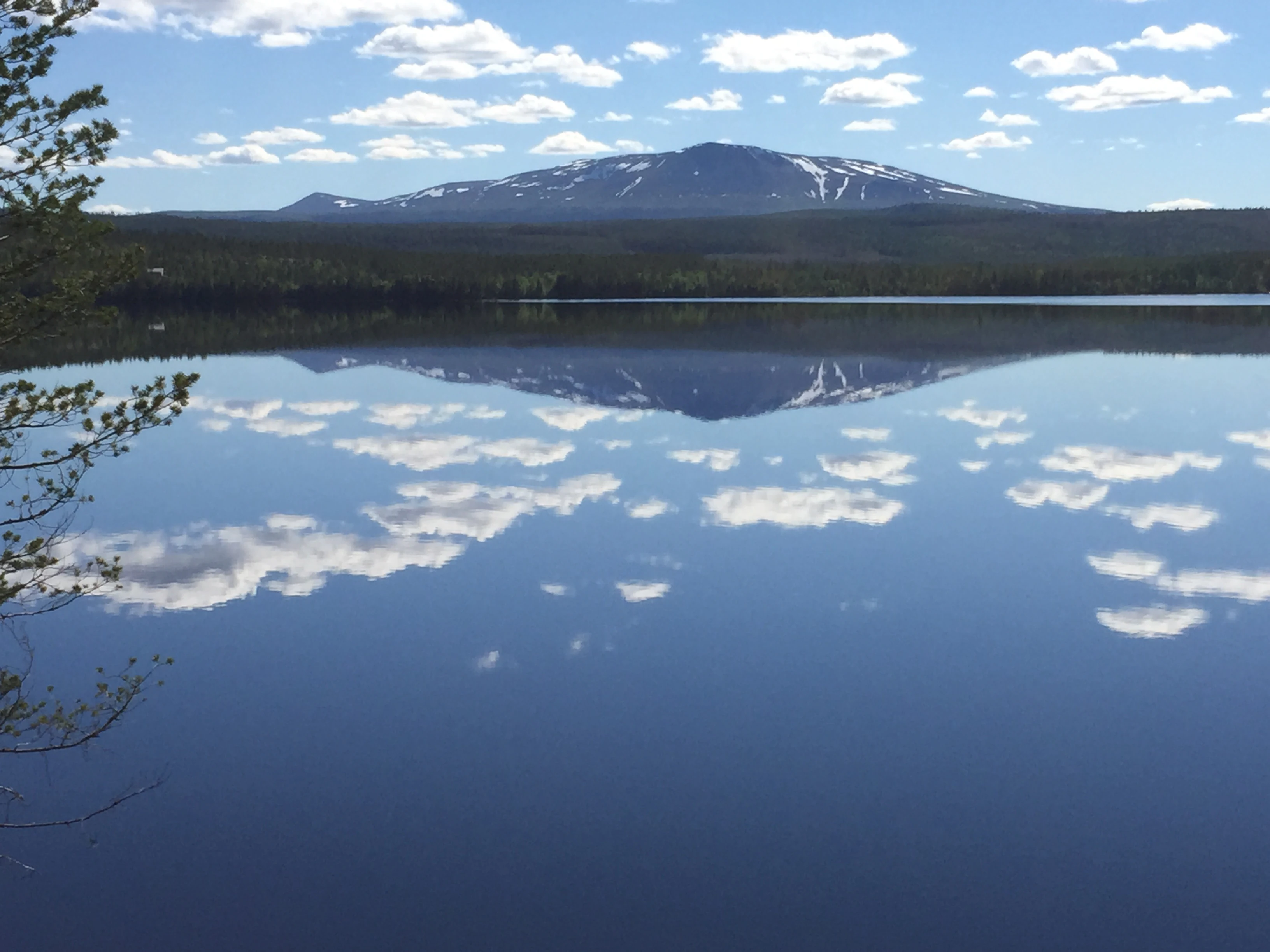 Calm lake reflecting mountain range and scattered clouds with tree branches on left