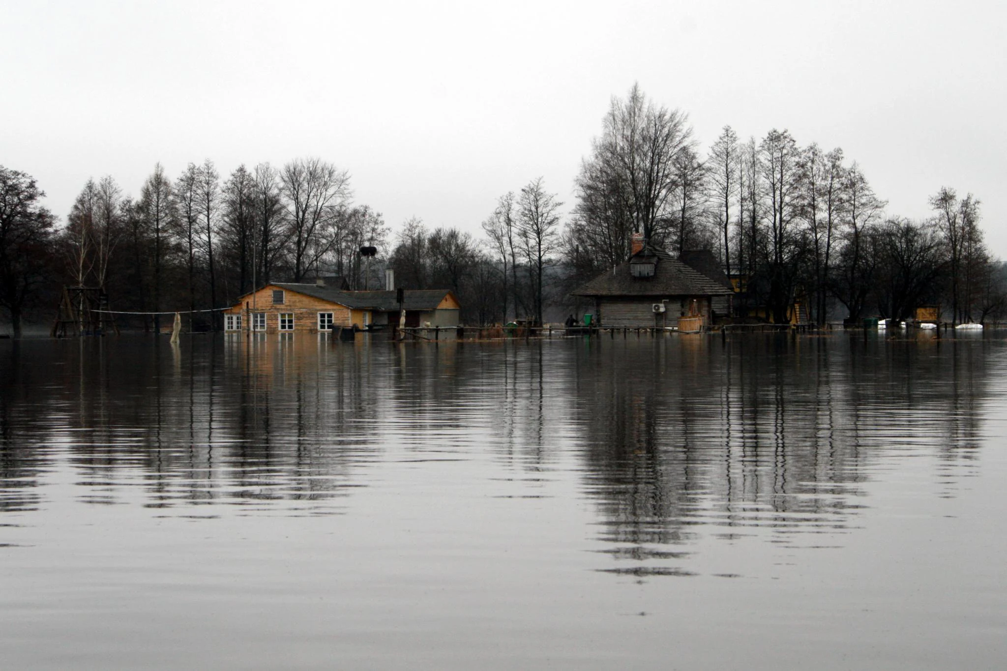 Waterlogged landscape with houses partially submerged in calm water, leafless trees in background, and overcast sky