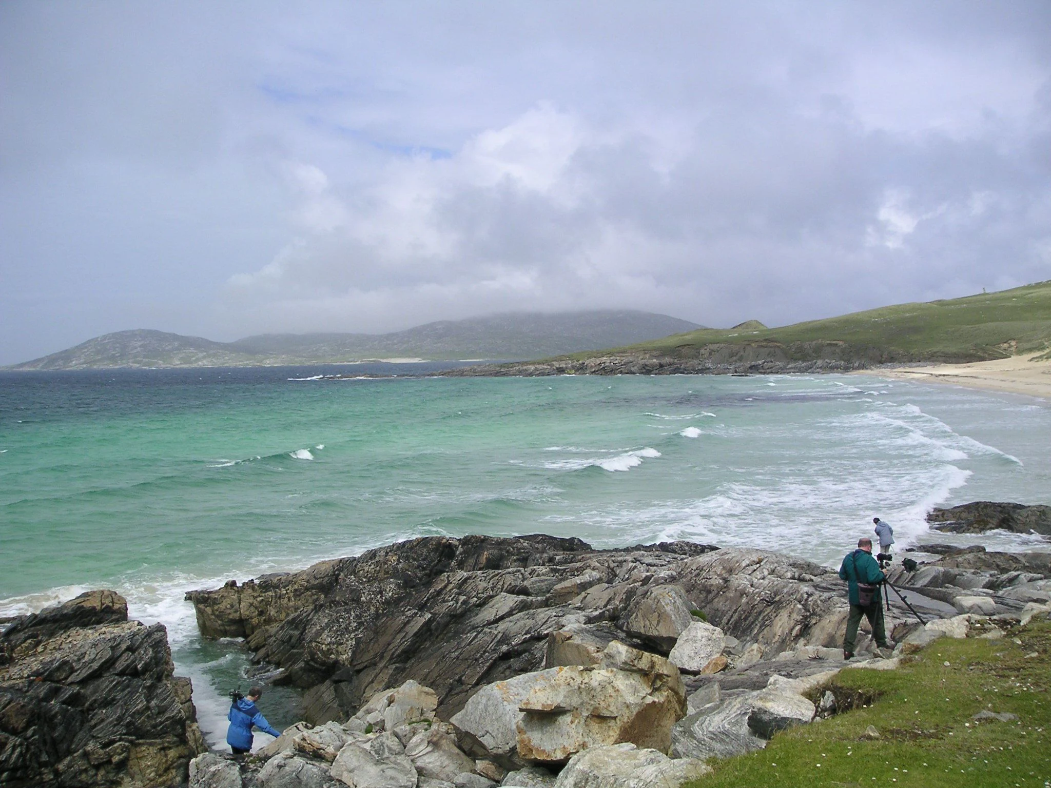 A wide coastal landscape in South Harris, Scotland, showing dark rocky foreground, turquoise sea with gentle waves, a curved sandy beach, and rolling green hills under a cloudy sky. Two people are standing on the rocks, one with a tripod.
