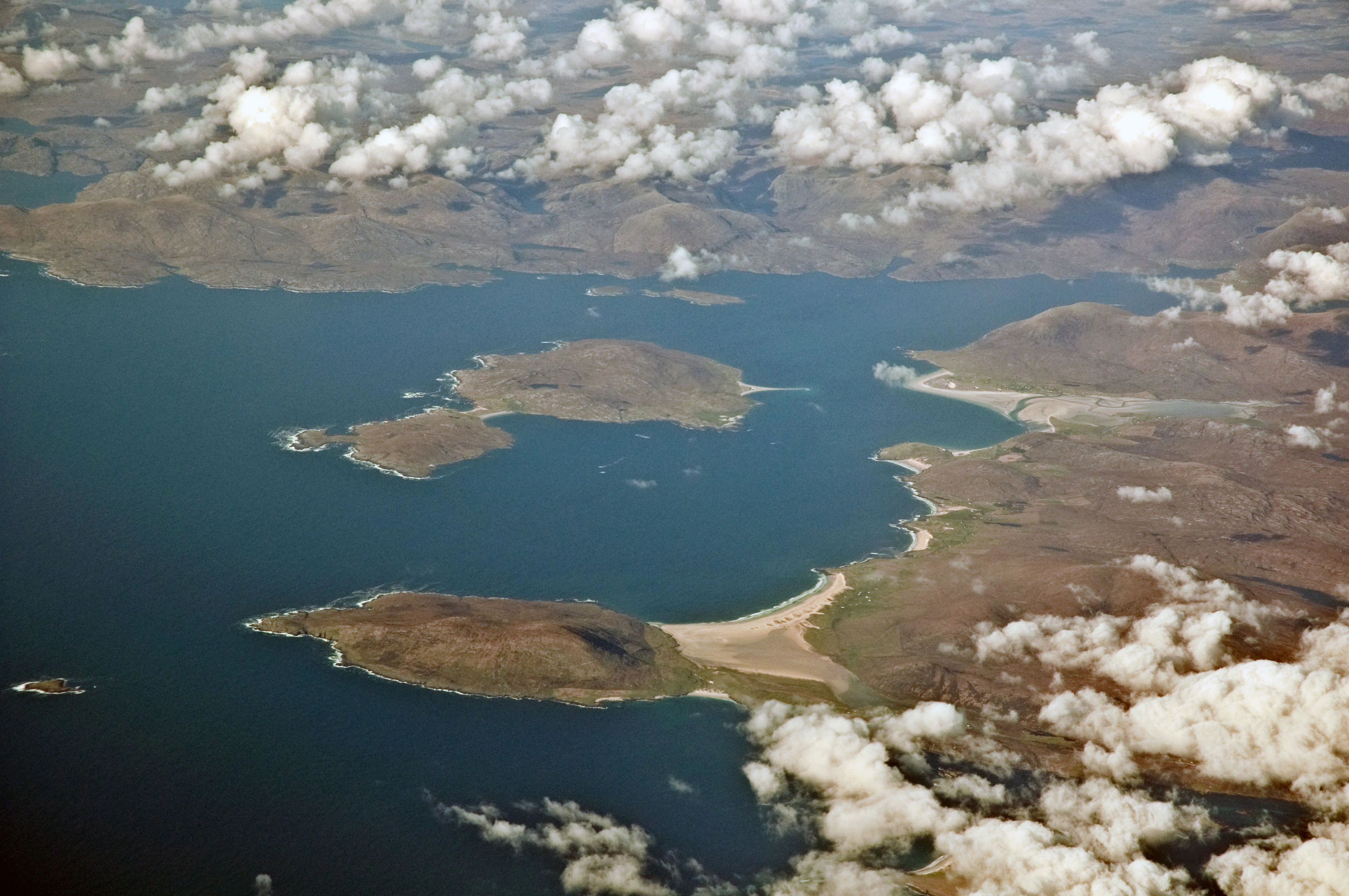 High-altitude aerial view of West Loch Tarbert in the Western Isles of Scotland, showing indented coastal islands, sandy shores, and surrounding rugged terrain partially obscured by scattered clouds.