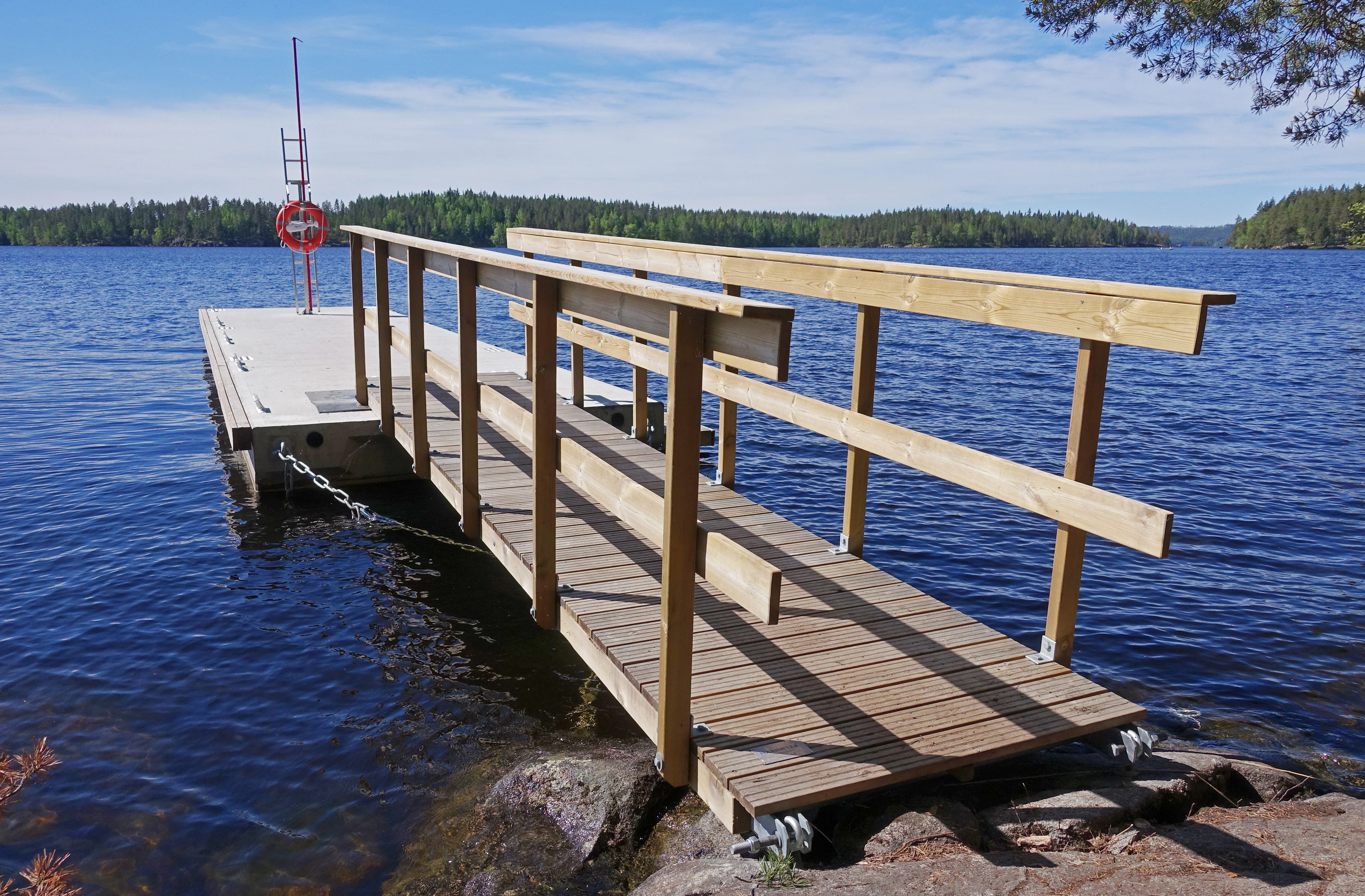 Wooden pier with railings extending from rocky shore into a lake, with forested land visible across the water