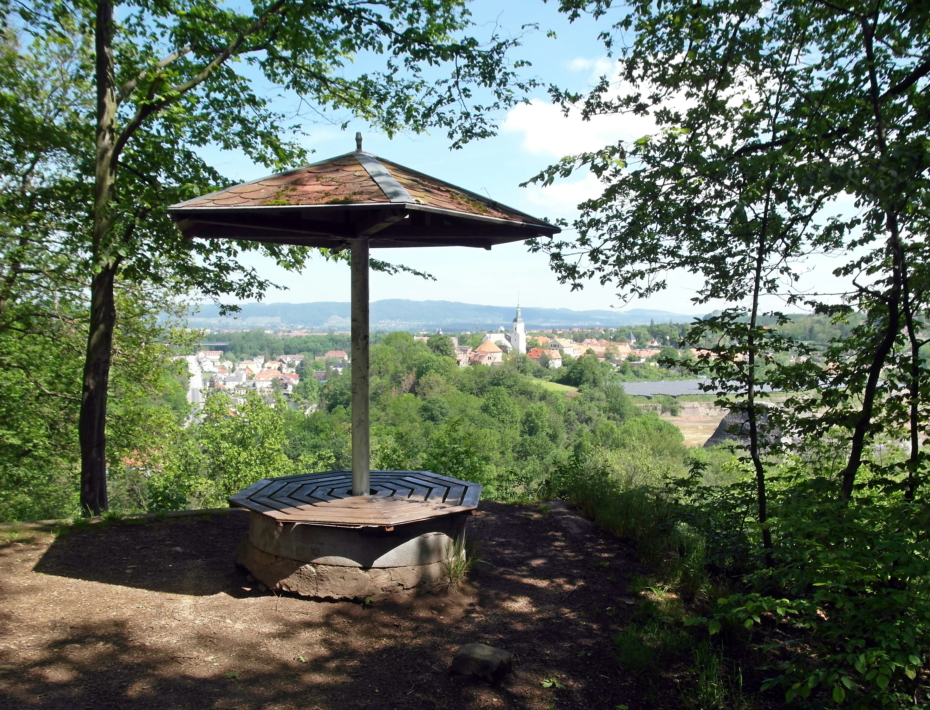 A wooden shelter with a central pillar and octagonal seating sits on a hillside overlooking a town with a prominent church steeple and distant hills.