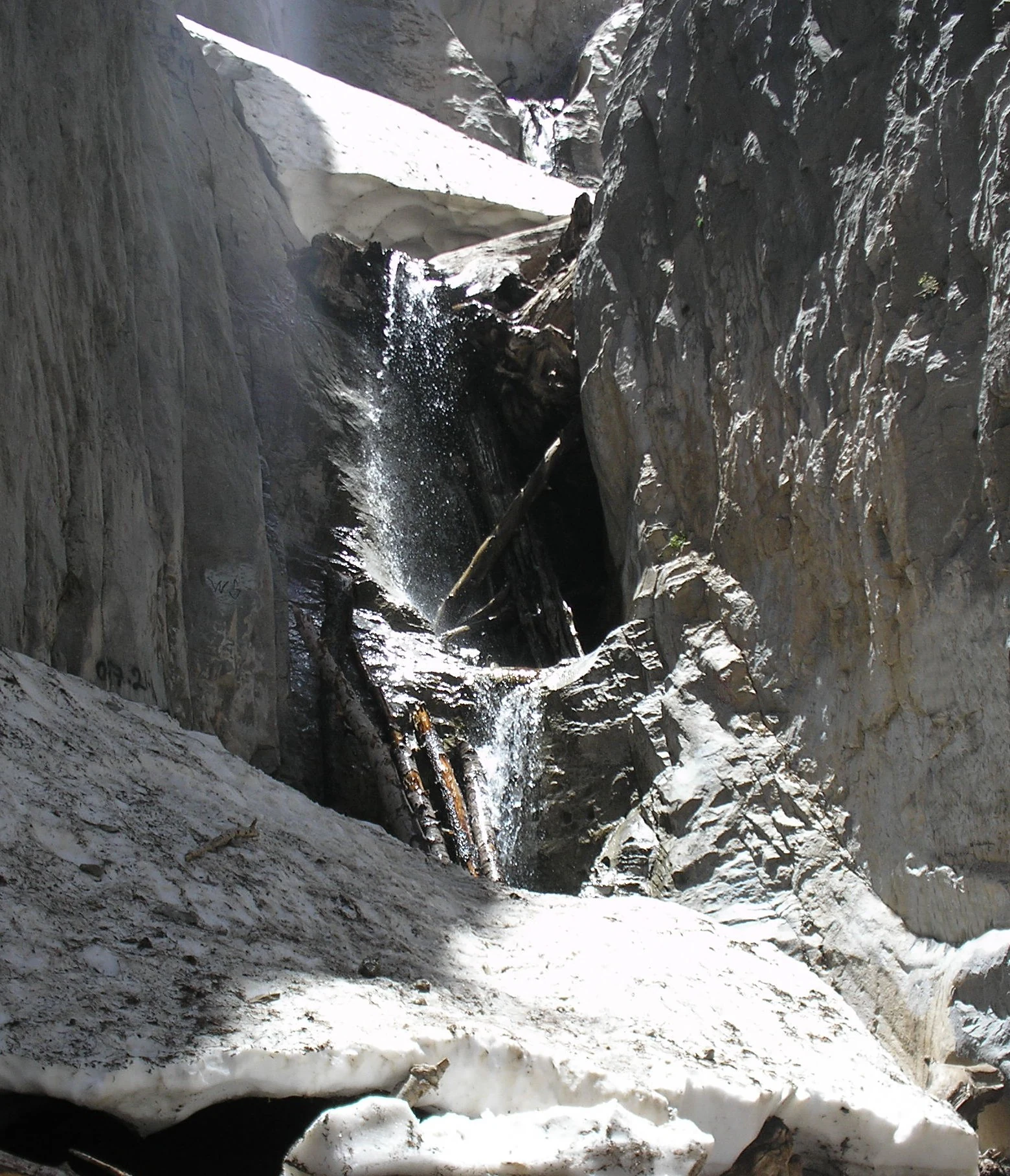 A small waterfall flowing between two high, steep rocky cliffs, with patches of snow remaining at the base and sides.