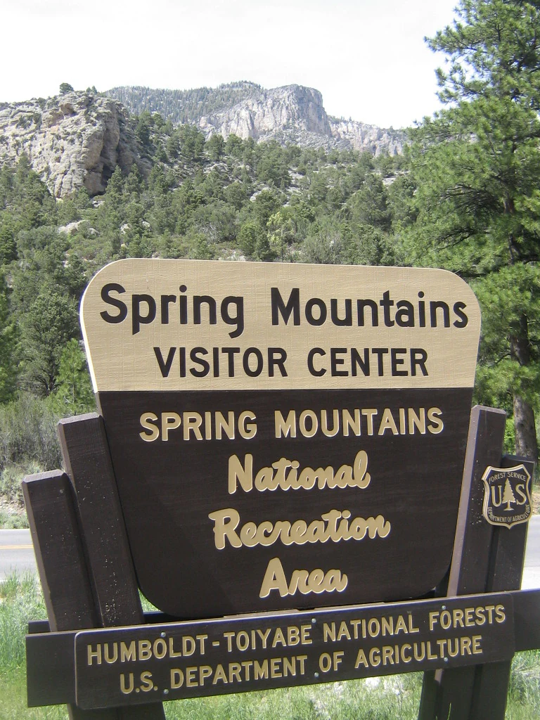 A wooden brown and beige entrance sign for the Spring Mountains Visitor Center, mounted on wooden posts with a mountain backdrop.