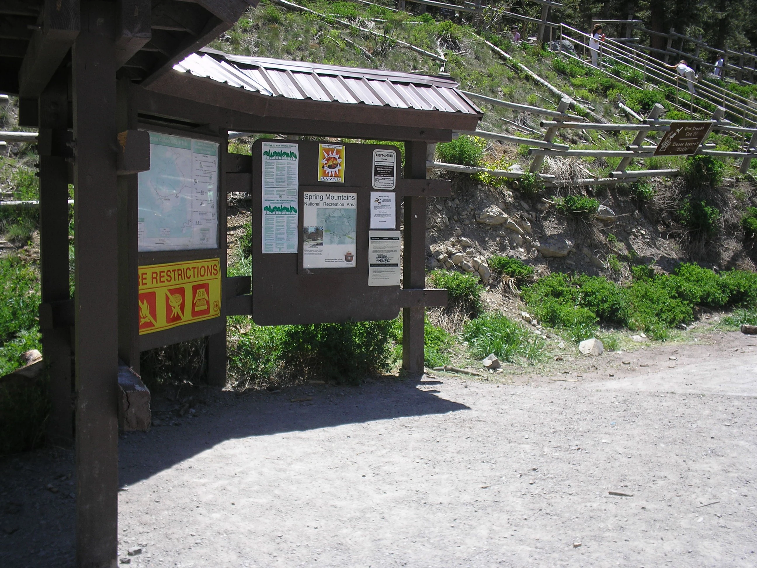 A wooden informational kiosk with multiple signs and maps stands near a dirt path at the base of a forested hillside with a wooden fence.