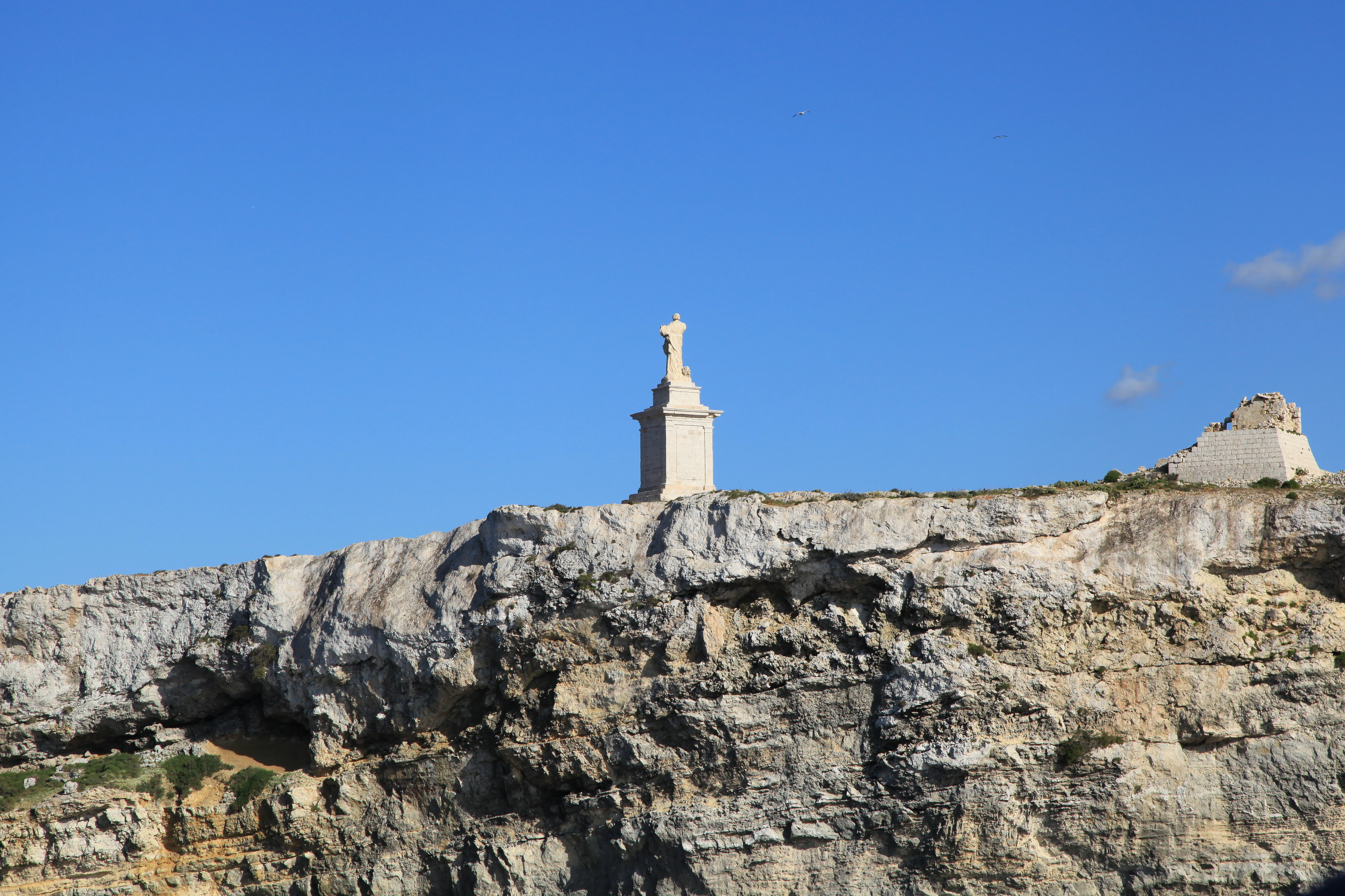 A white statue of St Paul stands atop a limestone cliff on a small island against a clear blue sky.