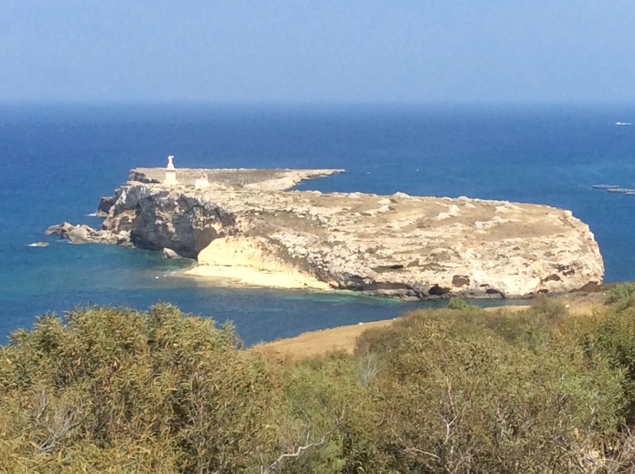 A wide angle view of the arid, rocky St Paul's Island surrounded by blue Mediterranean sea, seen from a distance across the water.