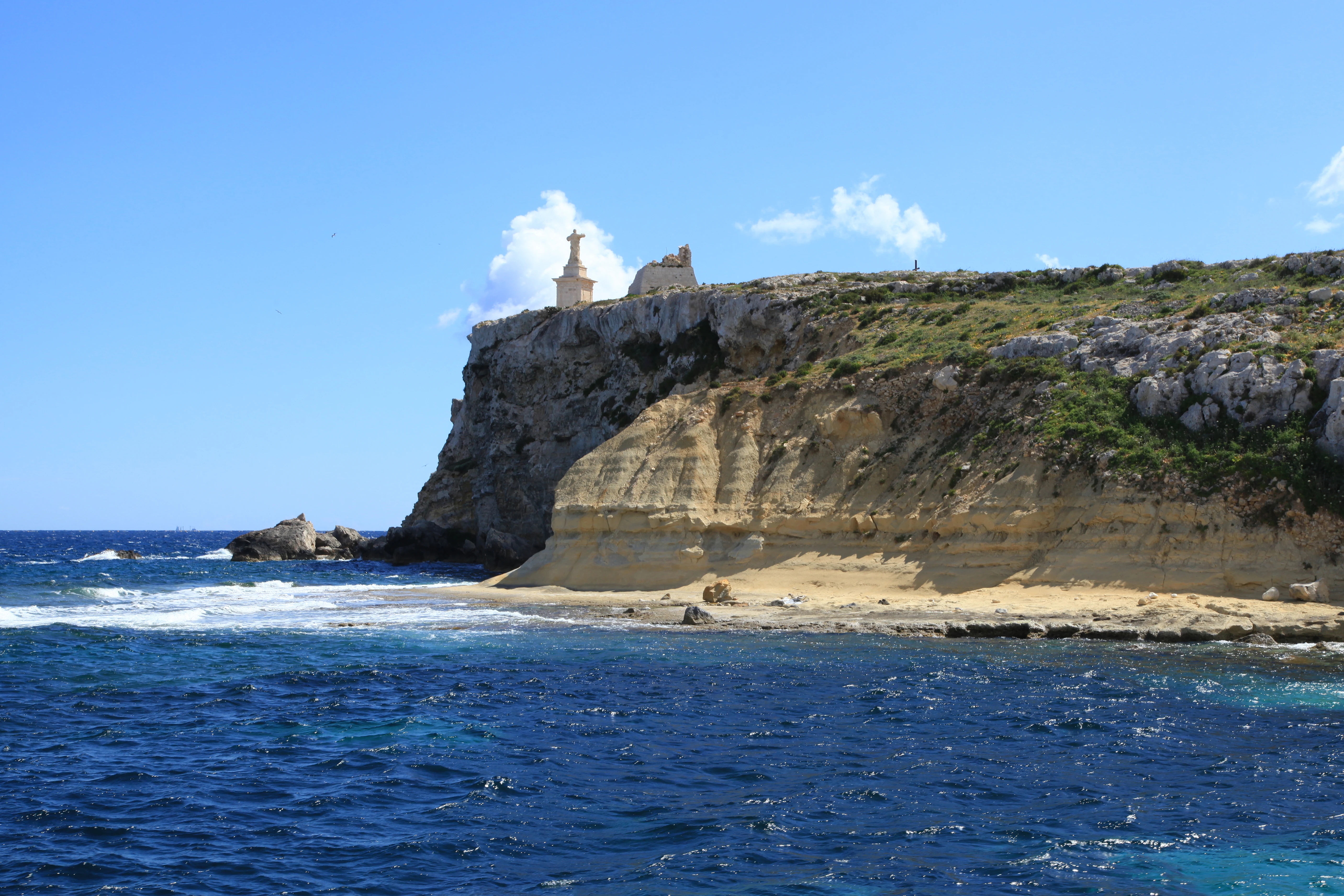 A sunny view from the sea looking at a limestone cliff on St Paul's Island, featuring a prominent white statue on the ridgeline.
