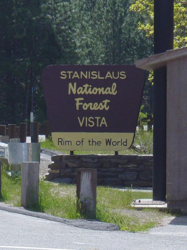 A brown and yellow official park sign reading Stanislaus National Forest VISTA Rim of the World, mounted on a stone base near a roadside.