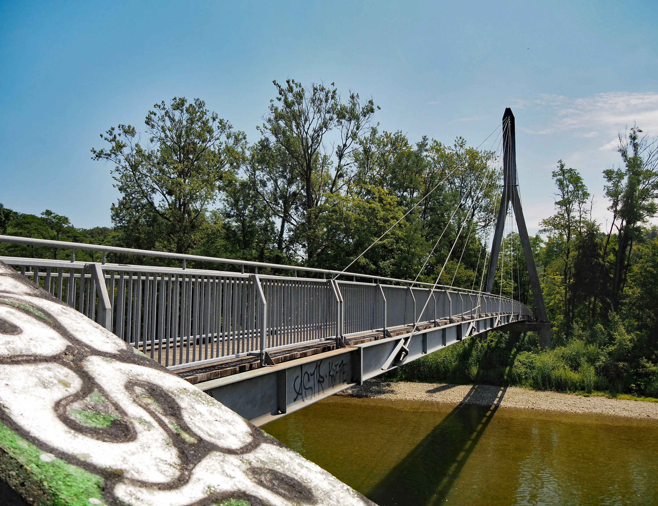 A modern grey metal suspension footbridge crossing a river; the bridge is angled from the foreground toward a tall, A-frame central pylon surrounded by green trees under a clear blue sky.