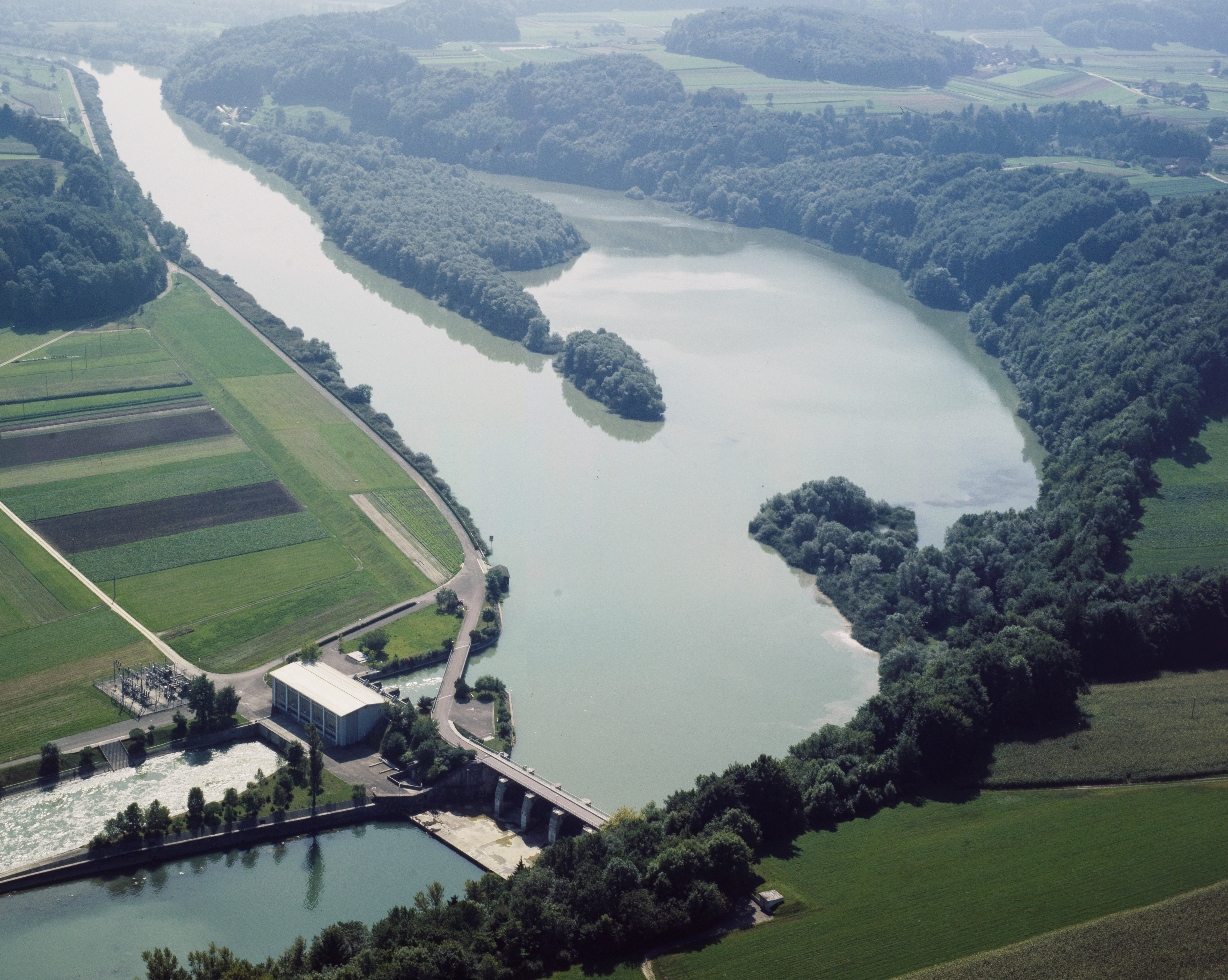 An aerial perspective of a reservoir on a river surrounded by forested banks and farmland, with a hydroelectric dam structure visible in the foreground.