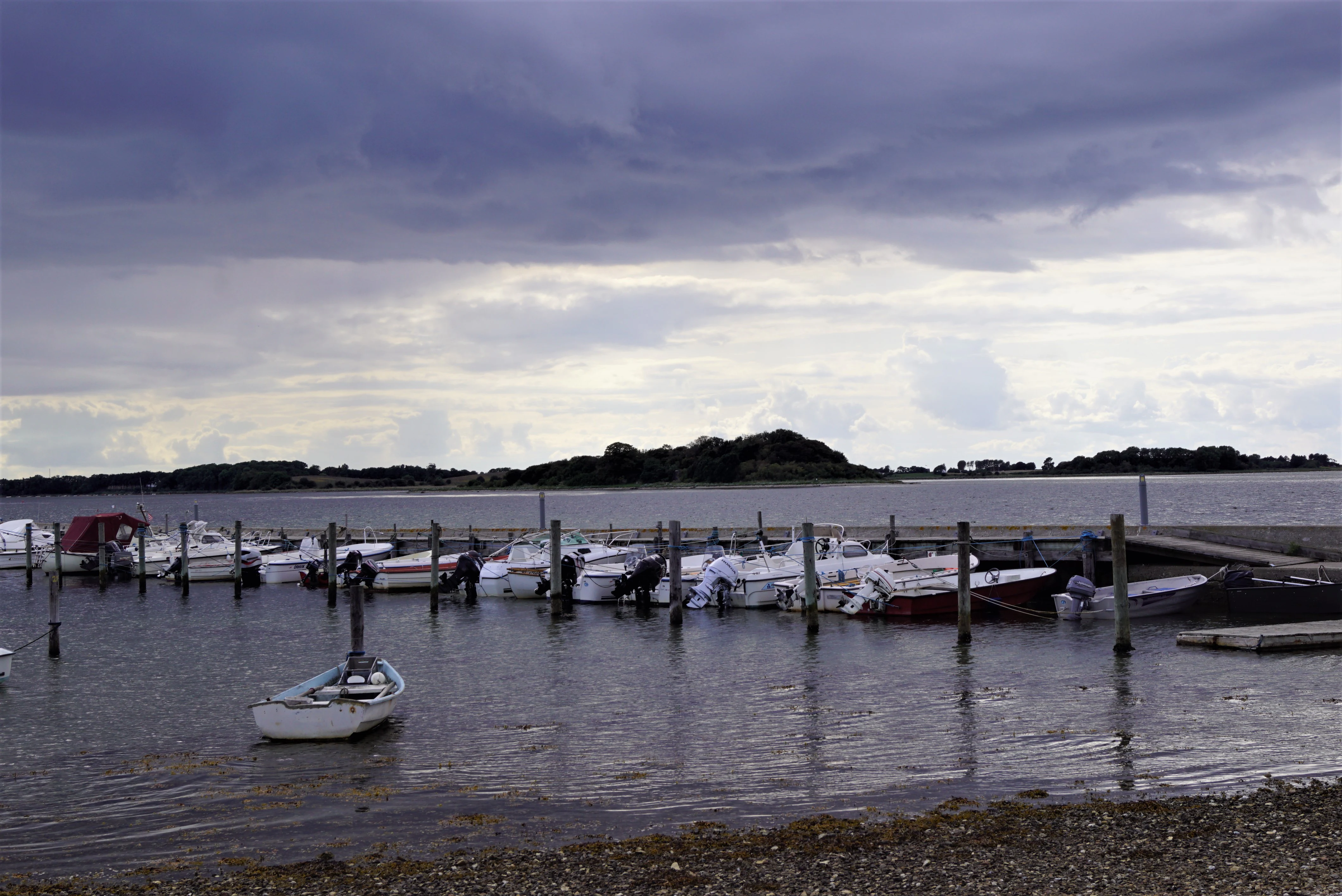 A line of small boats moored in a calm fjord harbor with a cloudy sky overhead.