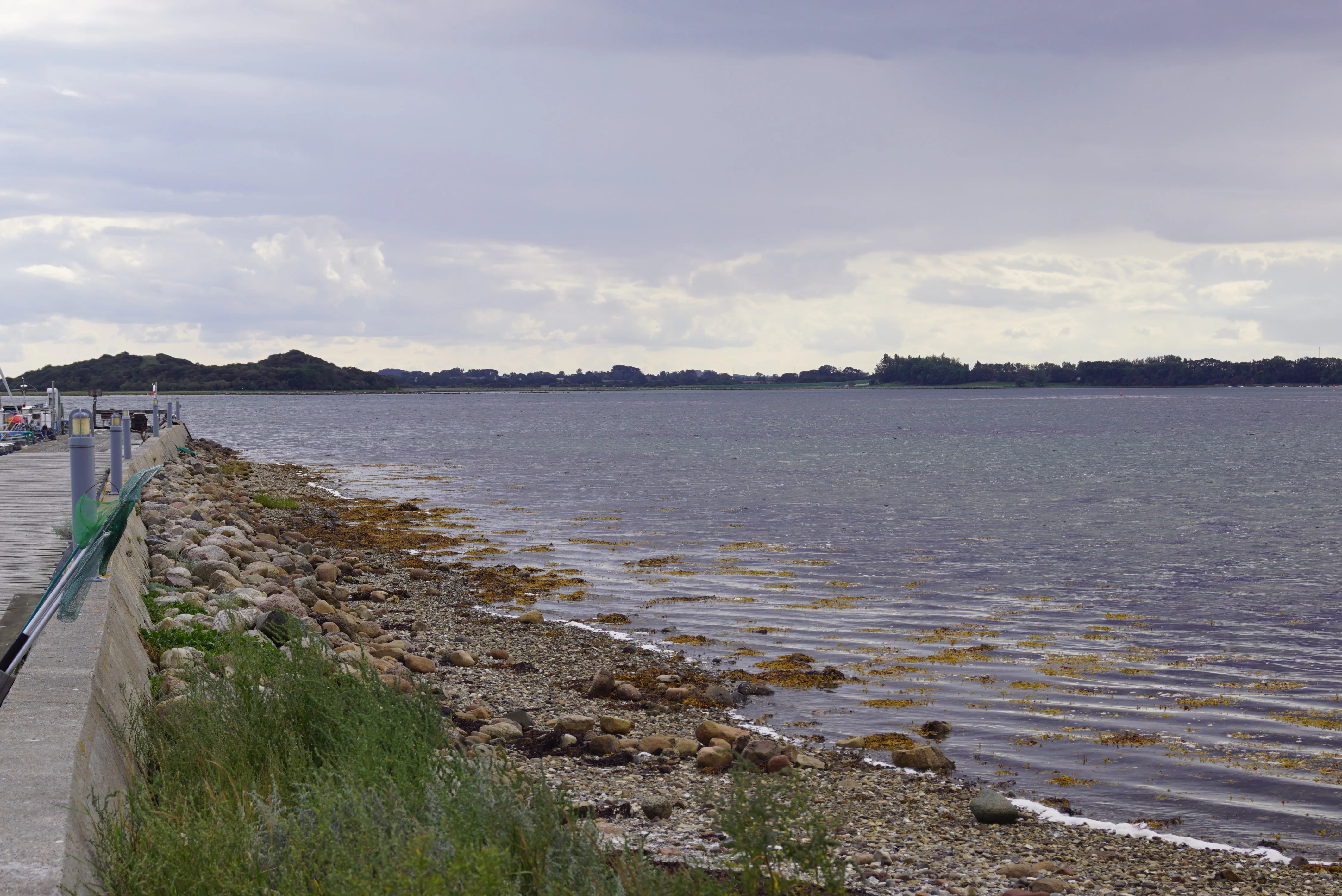 A coastal view of Stavns Fjord featuring a rocky shoreline, a concrete pier edge on the left, and distant low-lying land under a cloudy sky.