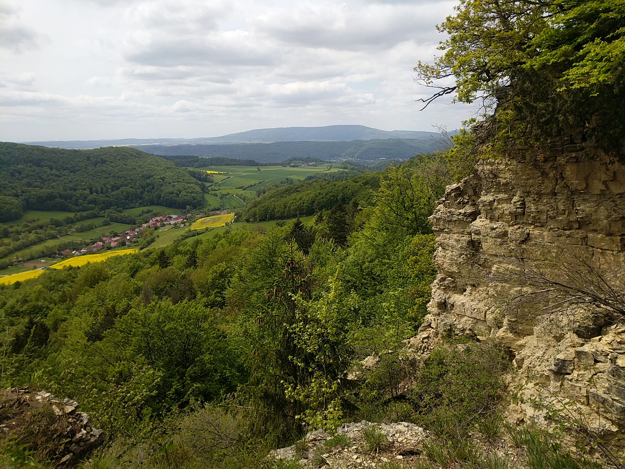 A high-angle landscape view showing a rugged limestone cliff face on the right foreground, overlooking a lush green valley with forests, yellow fields, a small village, and distant rolling hills under a cloudy sky.