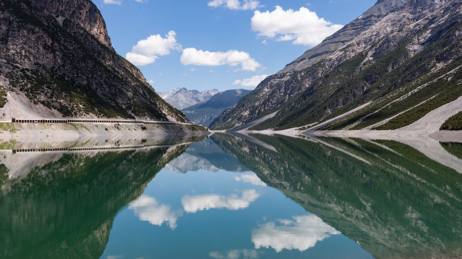 Calm lake reflecting steep rocky mountains and white clouds under a clear blue sky
