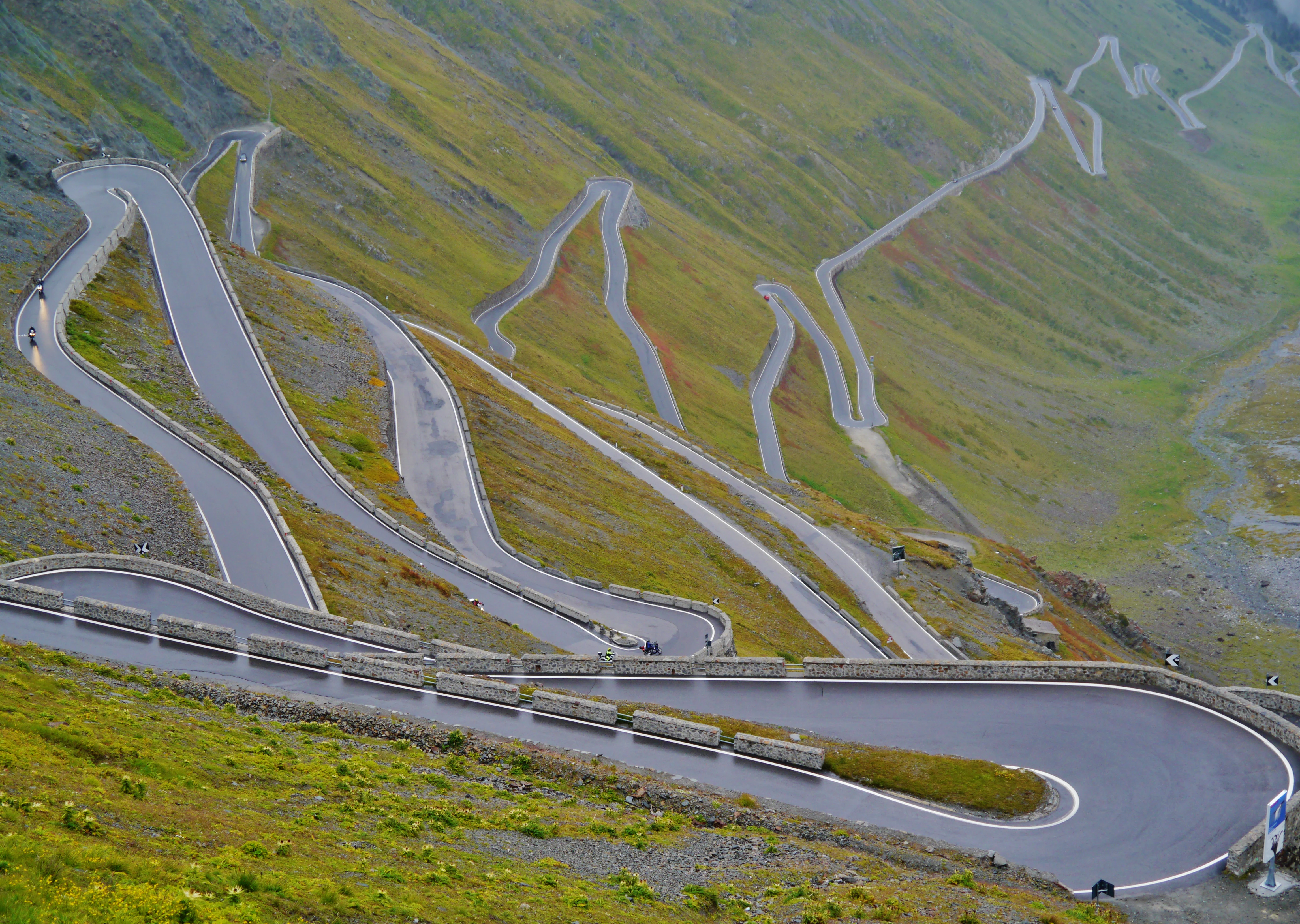 Winding mountain road with multiple hairpin turns on a steep slope, surrounded by alpine terrain and a blue sign