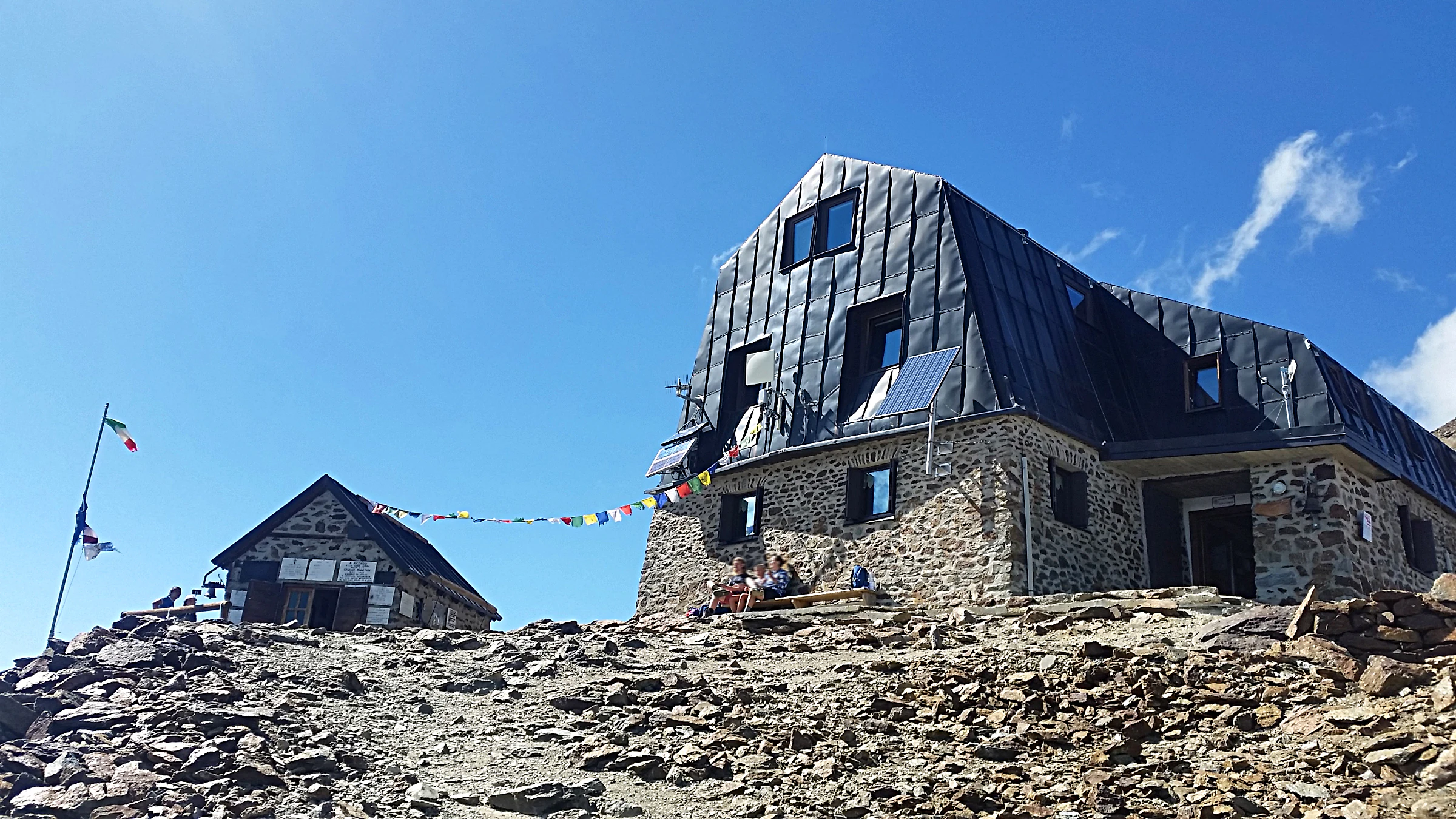 Stone mountain hut with dark roof and prayer flags strung across, rocky terrain, clear blue sky.