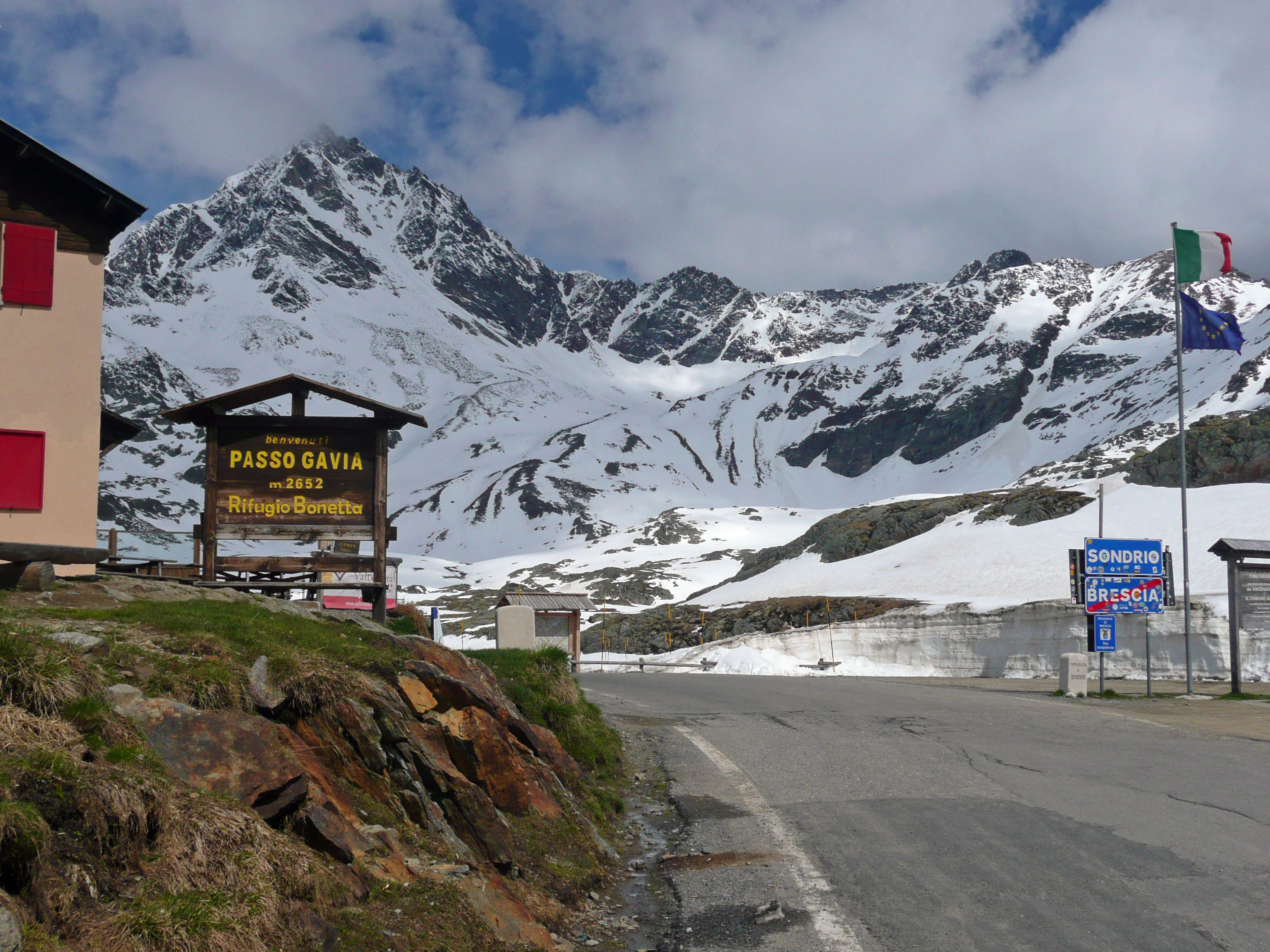 Sign indicating 'Passo Gavia' and 'Rifugio Bonetta' at a mountain pass with snow-covered peaks, road, and Italian and regional flags