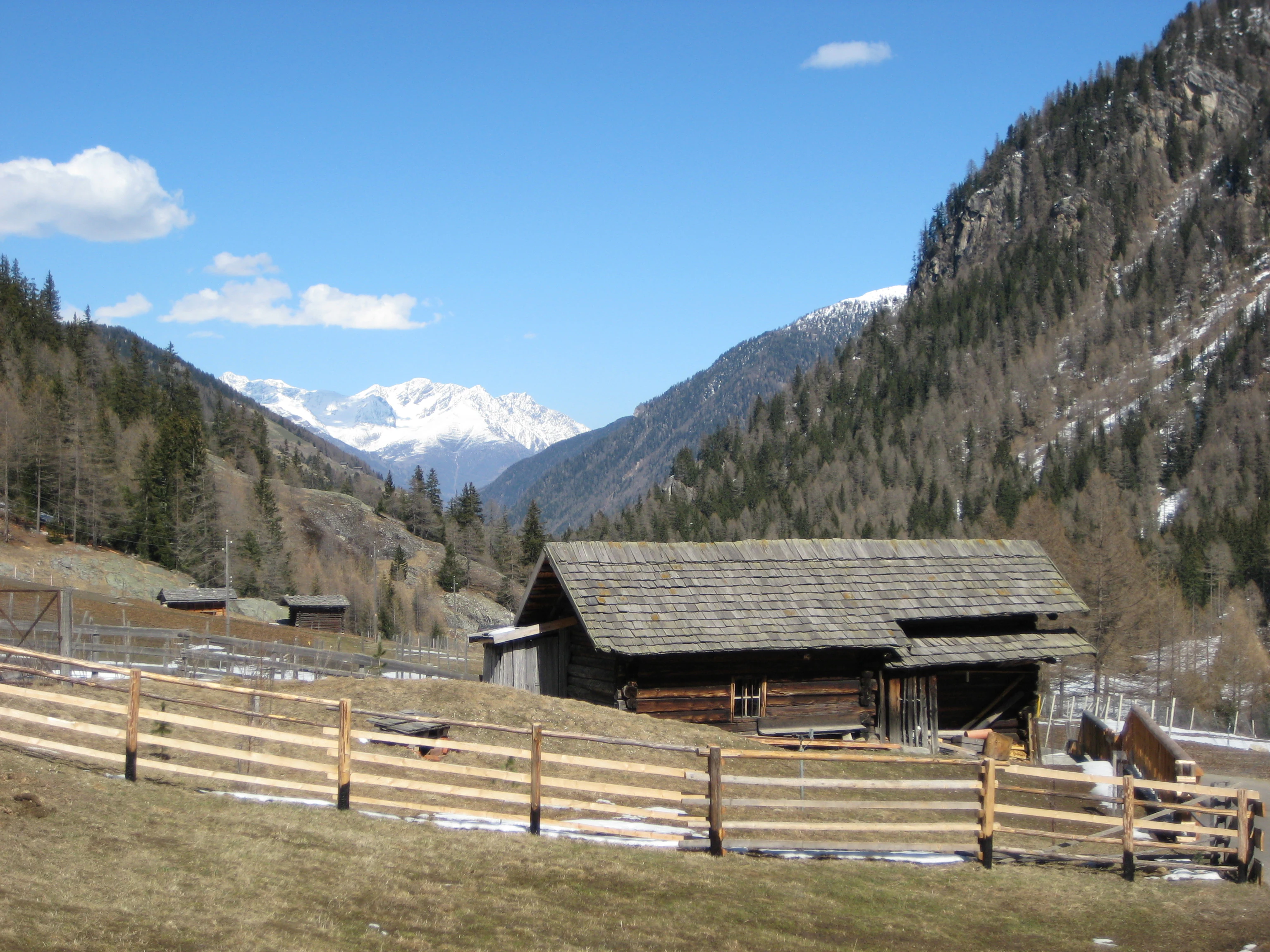 Wooden cabin-like structure with fence in mountain valley, snow-capped peaks in background
