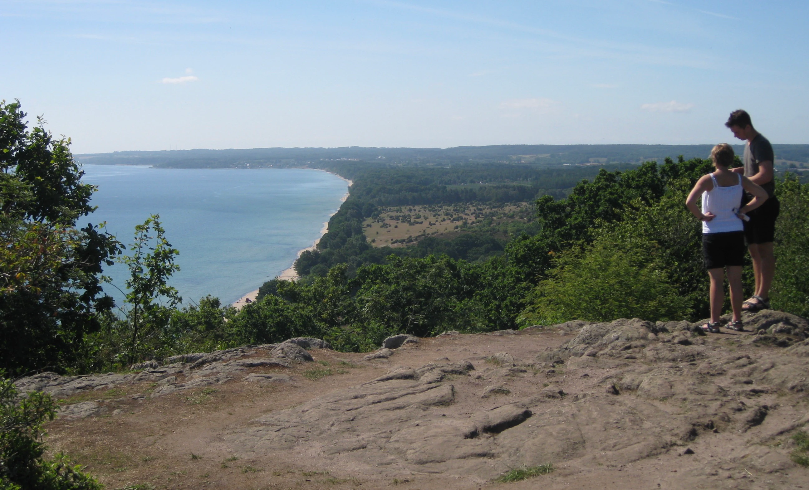 Two people standing on a rocky hilltop with a coastal landscape view including a sandy beach, forest, and blue sea under a clear sky.