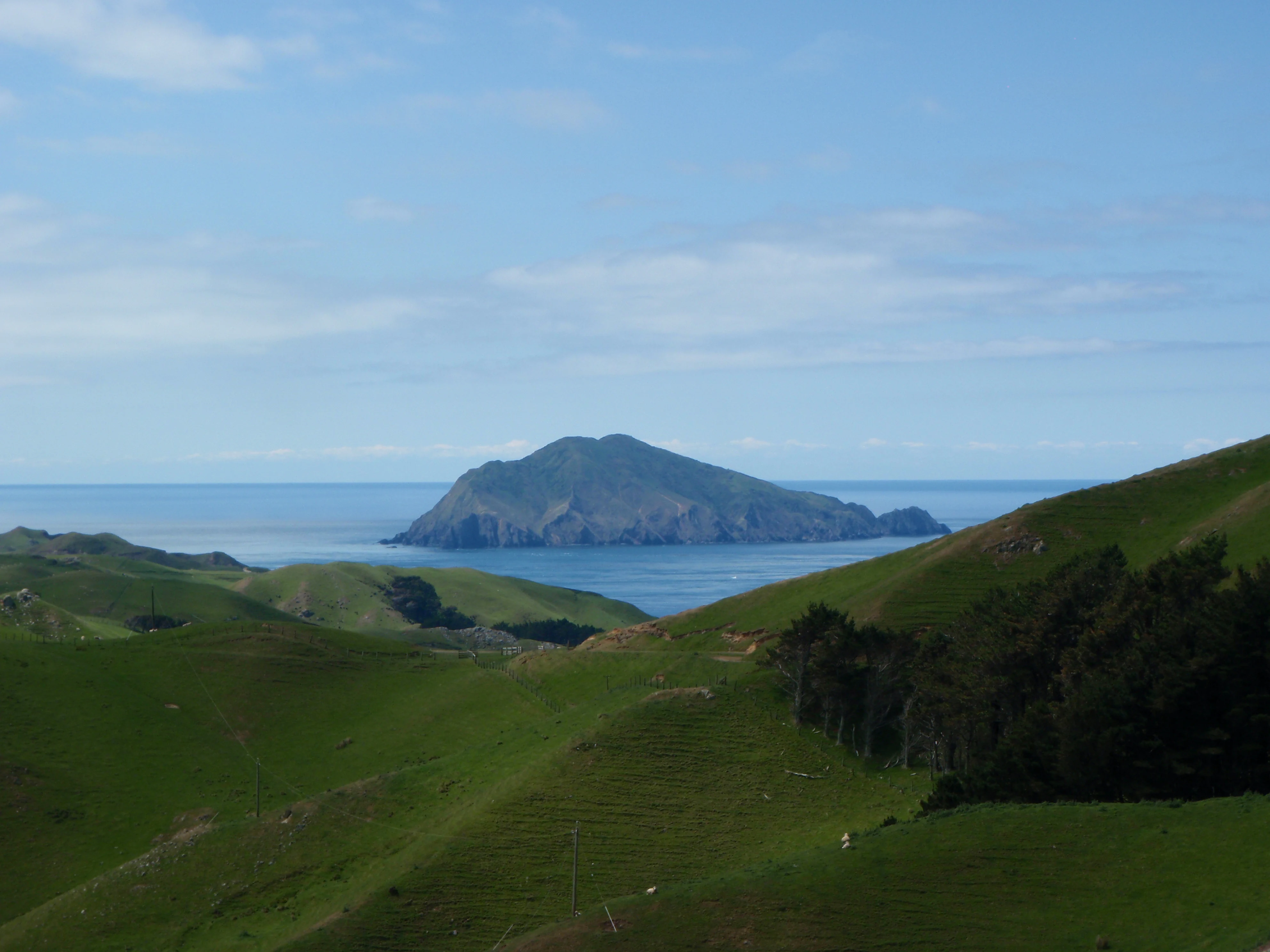 Una vista lejana de una isla escarpada y cubierta de hierba rodeada de mar azul, vista desde colinas verdes onduladas en primer plano.