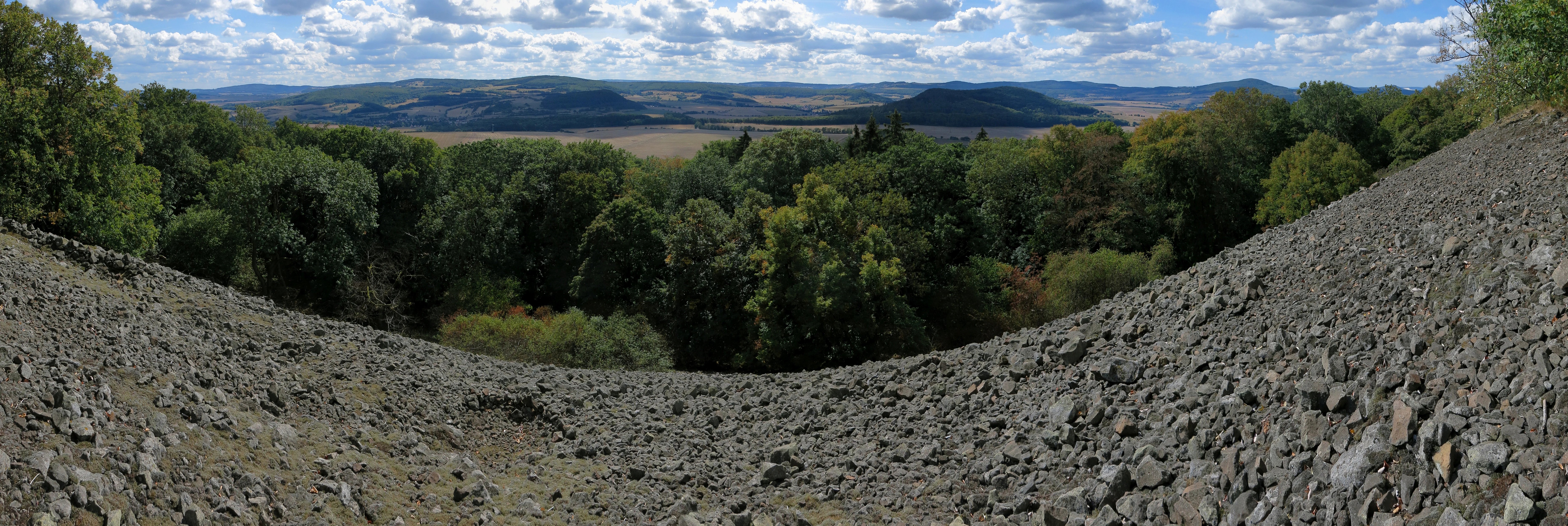 A wide panoramic photograph showing a rocky basalt talus slope in the foreground leading down to a dense forest canopy and distant rolling hills under a blue sky with scattered clouds.