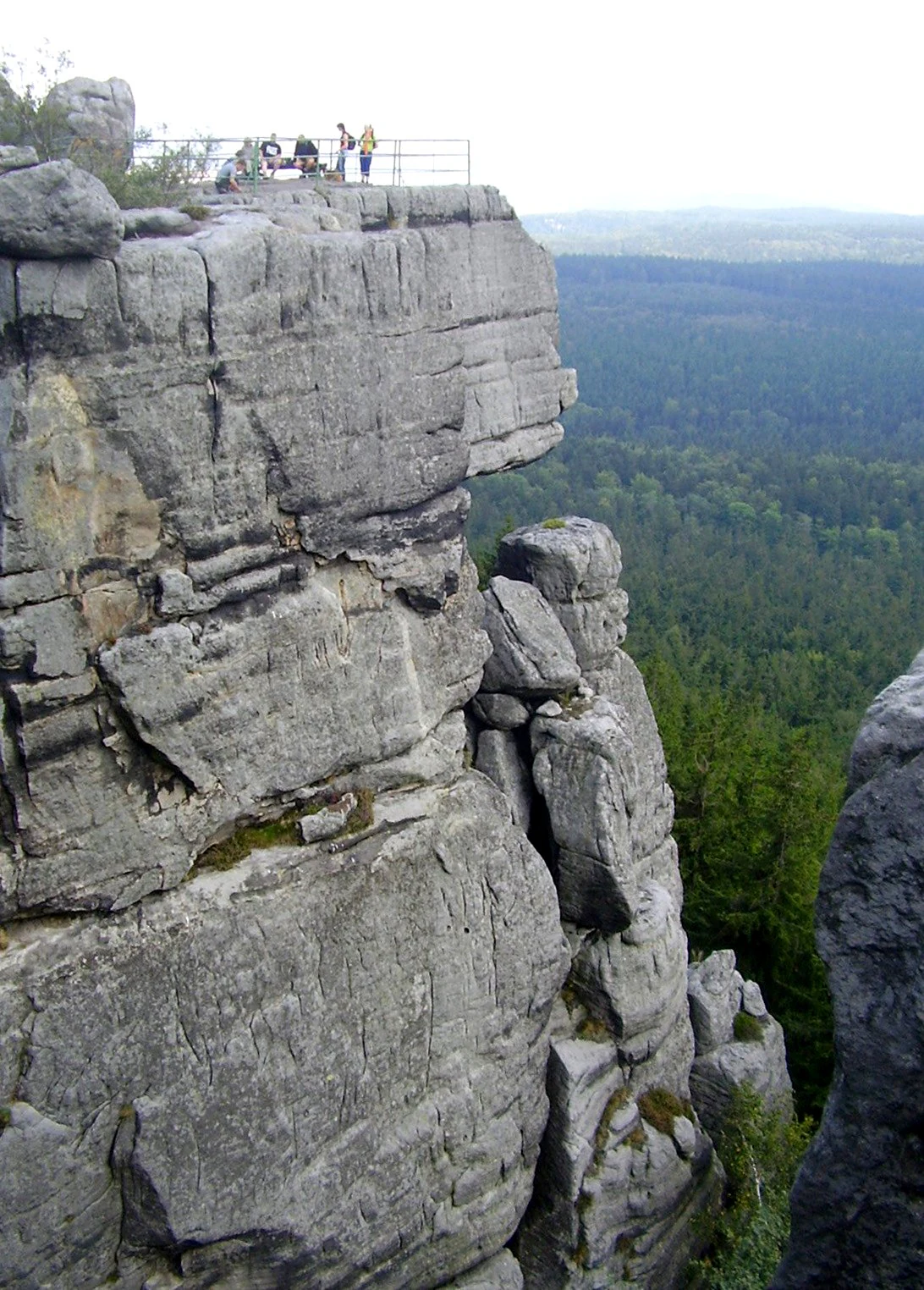 Rocky viewpoint terrace with several people standing on the edge, overlooking a dense forest below