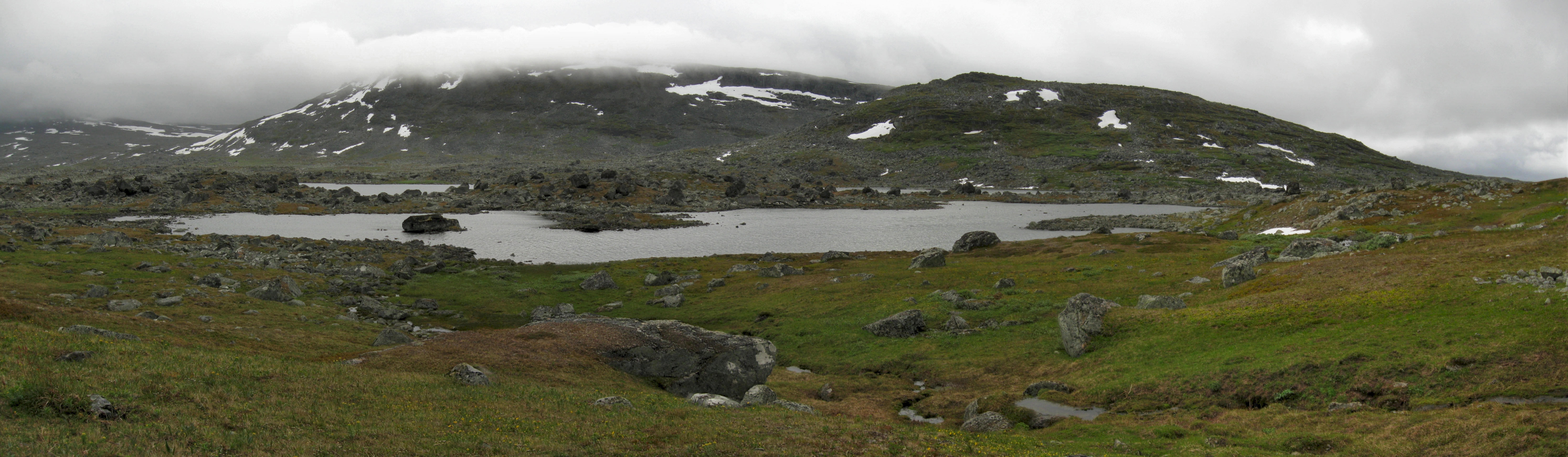 Panoramic view of Lake Nuortap Adtji surrounded by grassy terrain and rocky outcrops, with snow-capped mountains under a cloudy sky