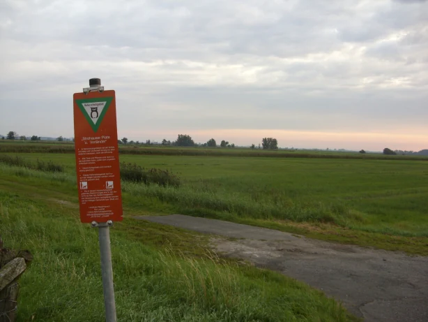 An orange nature reserve sign stands on a metal post at the edge of a grassy meadow and a small paved path under an overcast sky.
