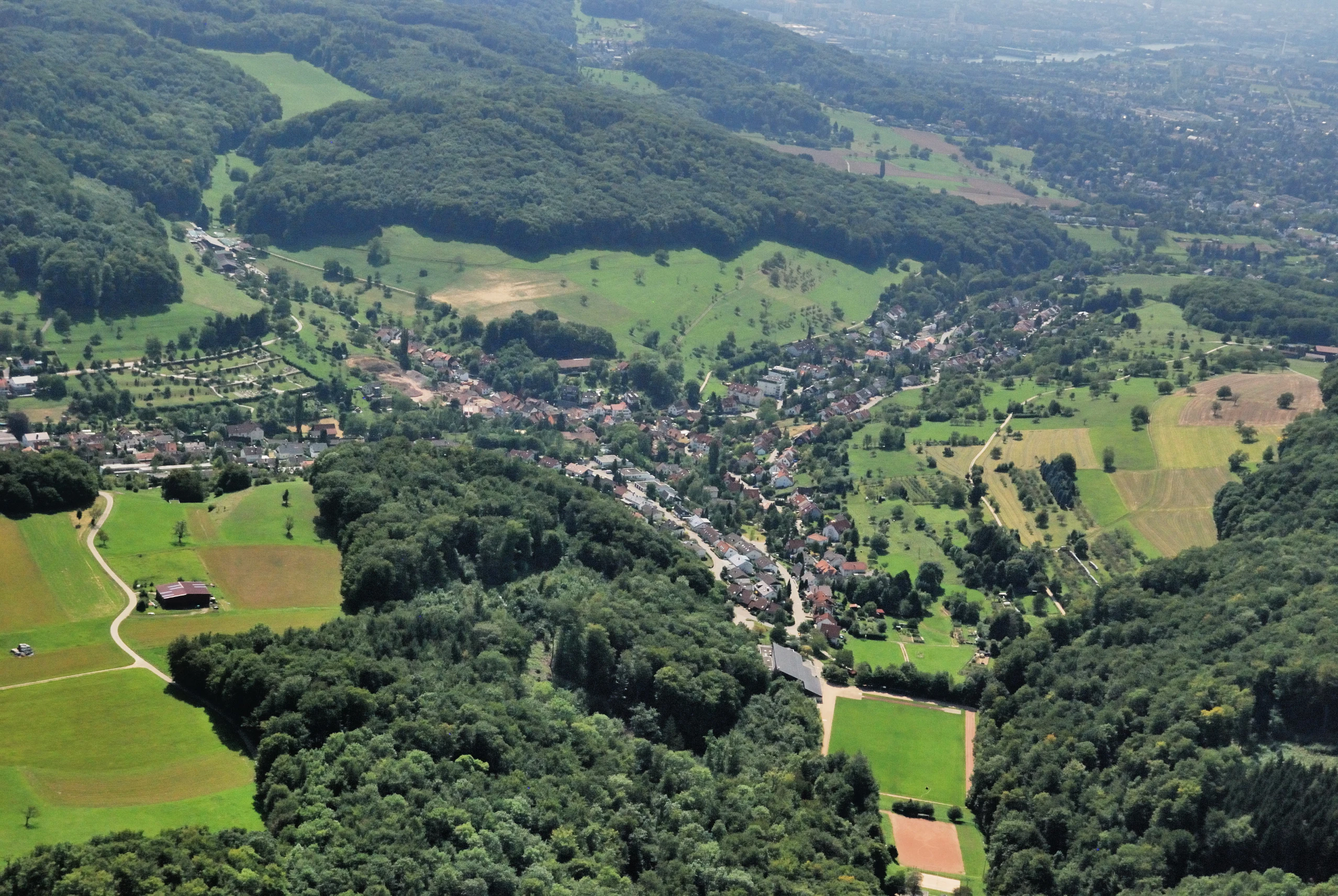 High-angle aerial view of a village surrounded by dense green forests and agricultural fields in a hilly landscape.