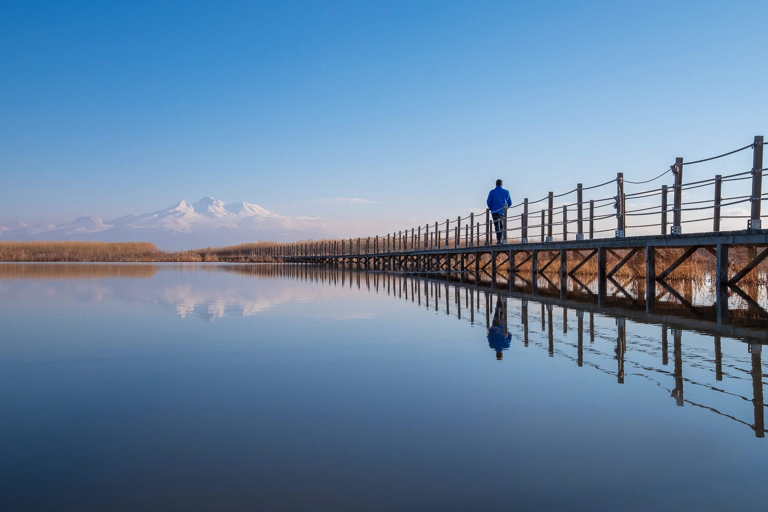 Wooden bridge over calm water with a person walking on it, reflection visible in water, snow-capped mountains in the background