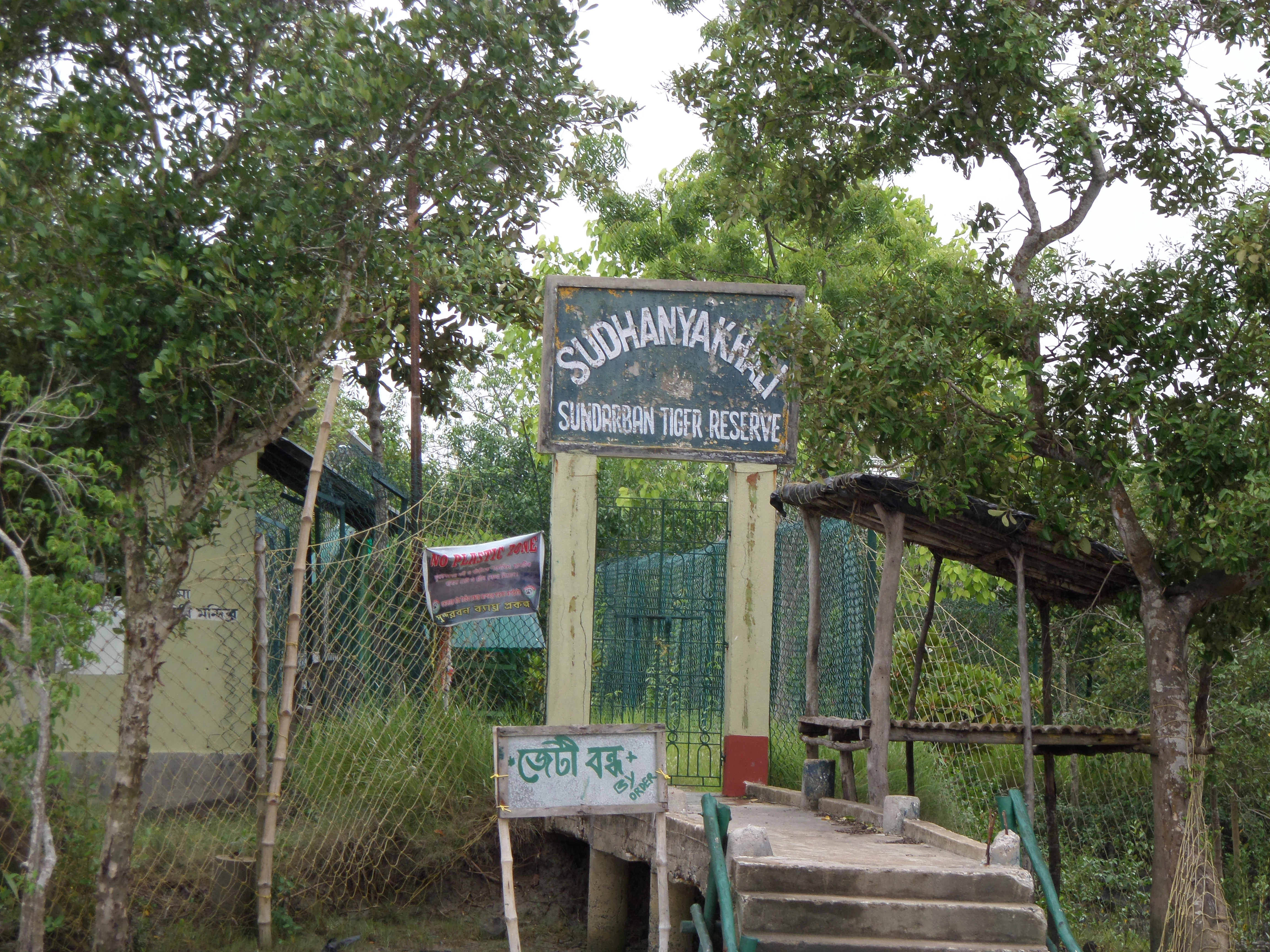 Cartel de entrada del área de Sudhanyakhali de la Reserva de Tigres de Sundarbans con texto en bengalí debajo, rodeado de árboles y vegetación