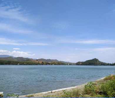 Una toma amplia a la altura de los ojos de un gran lago con agua azul y tranquila, rodeado de colinas onduladas bajo un cielo azul brillante con nubes dispersas.