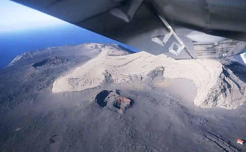 An aerial view from an airplane window showing the dark, volcanic landscape of Surtsey island, featuring a prominent crater and light-colored deposits against the ocean backdrop.