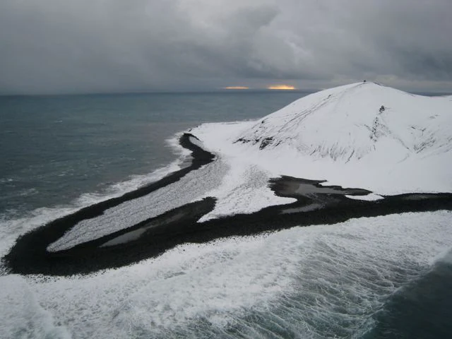 An aerial view of the Surtsey island shoreline, showing a black volcanic sand spit extending into the ocean, partially covered in snow under a cloudy, overcast sky.