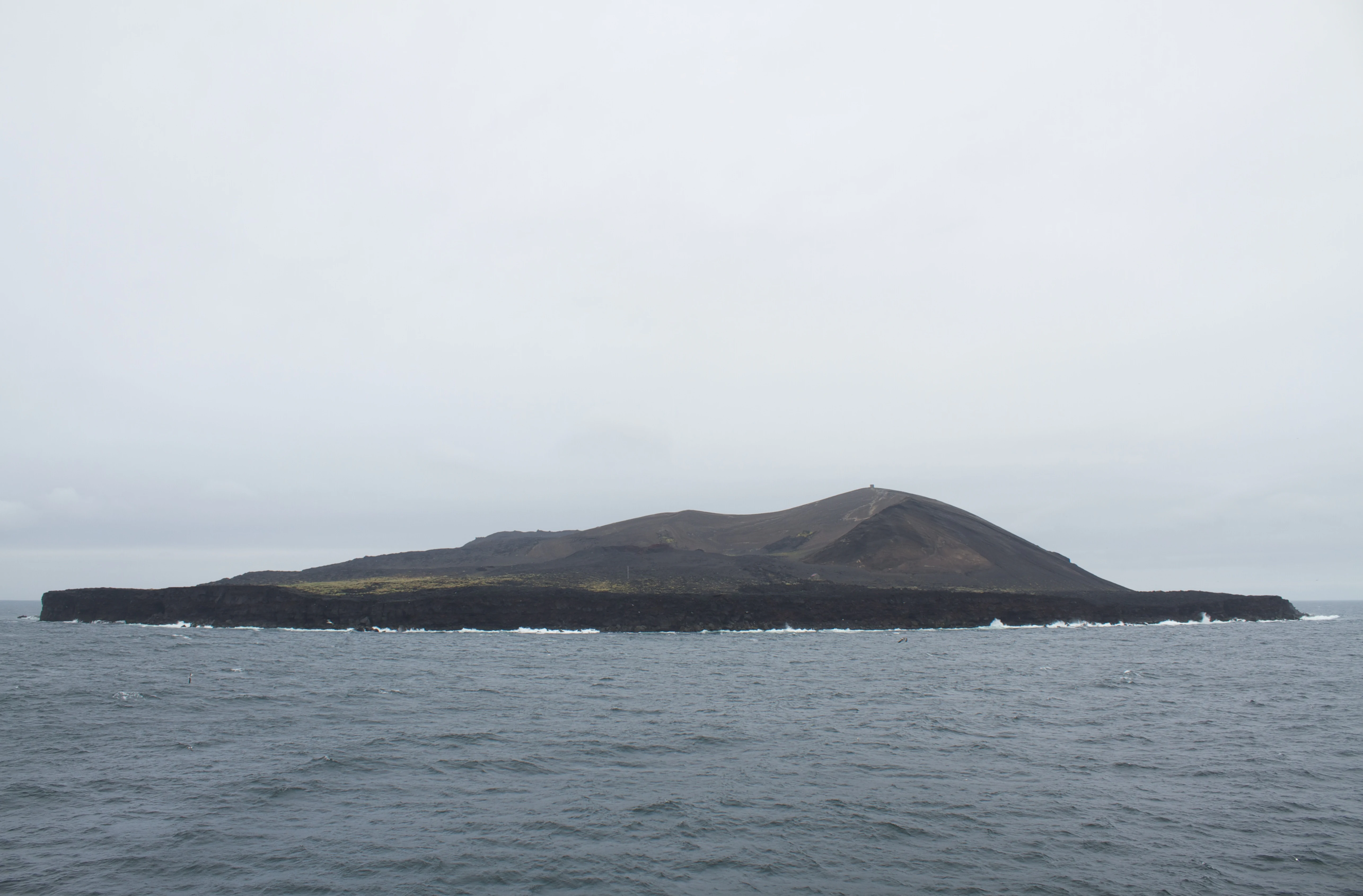 A low, dark, volcanic island rises from the gray sea under an overcast sky.